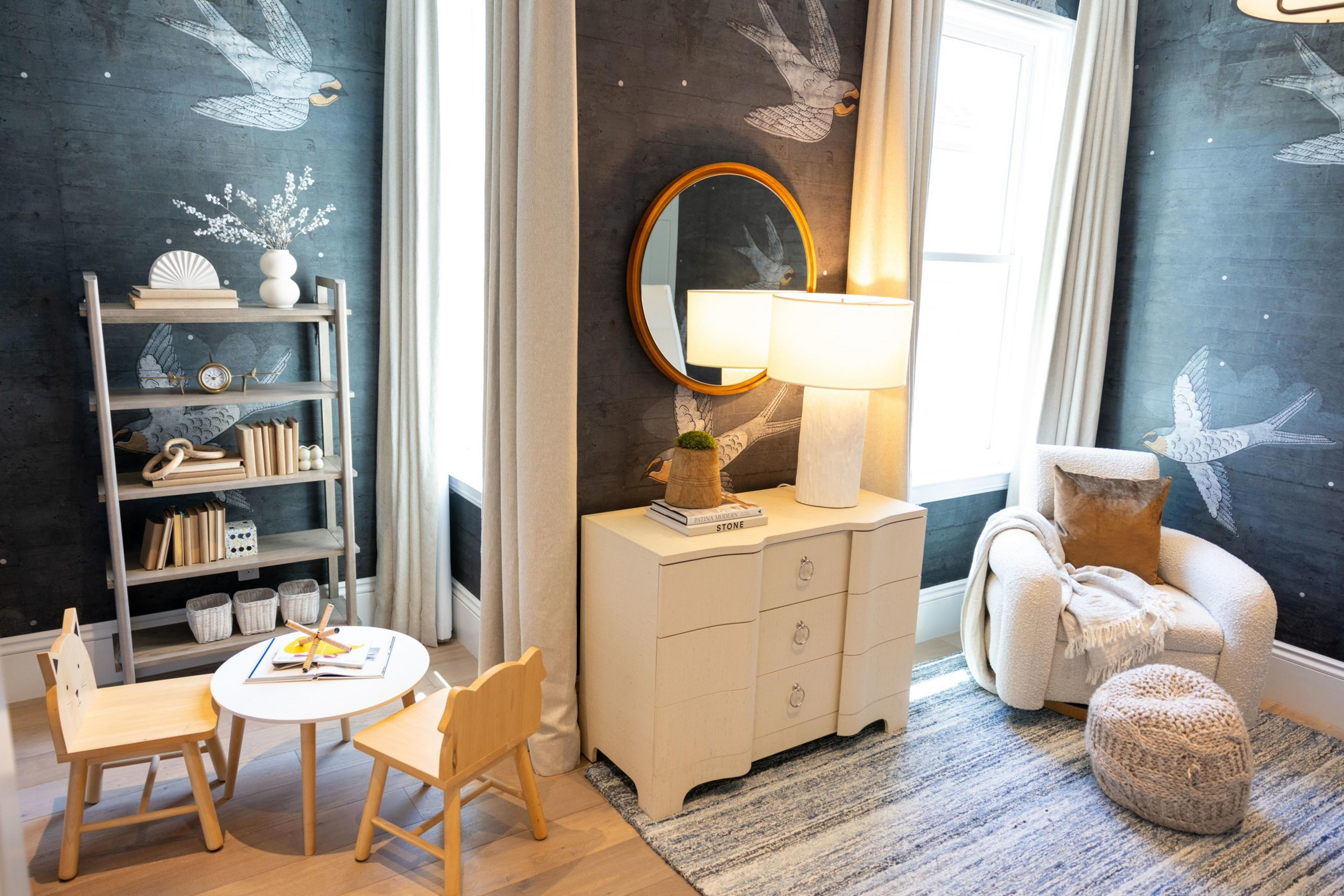 A cozy, stylish room features a small wooden table with children's chairs, a dresser with a lamp, a cushioned chair with a throw, a bookshelf, and walls decorated with bird murals.