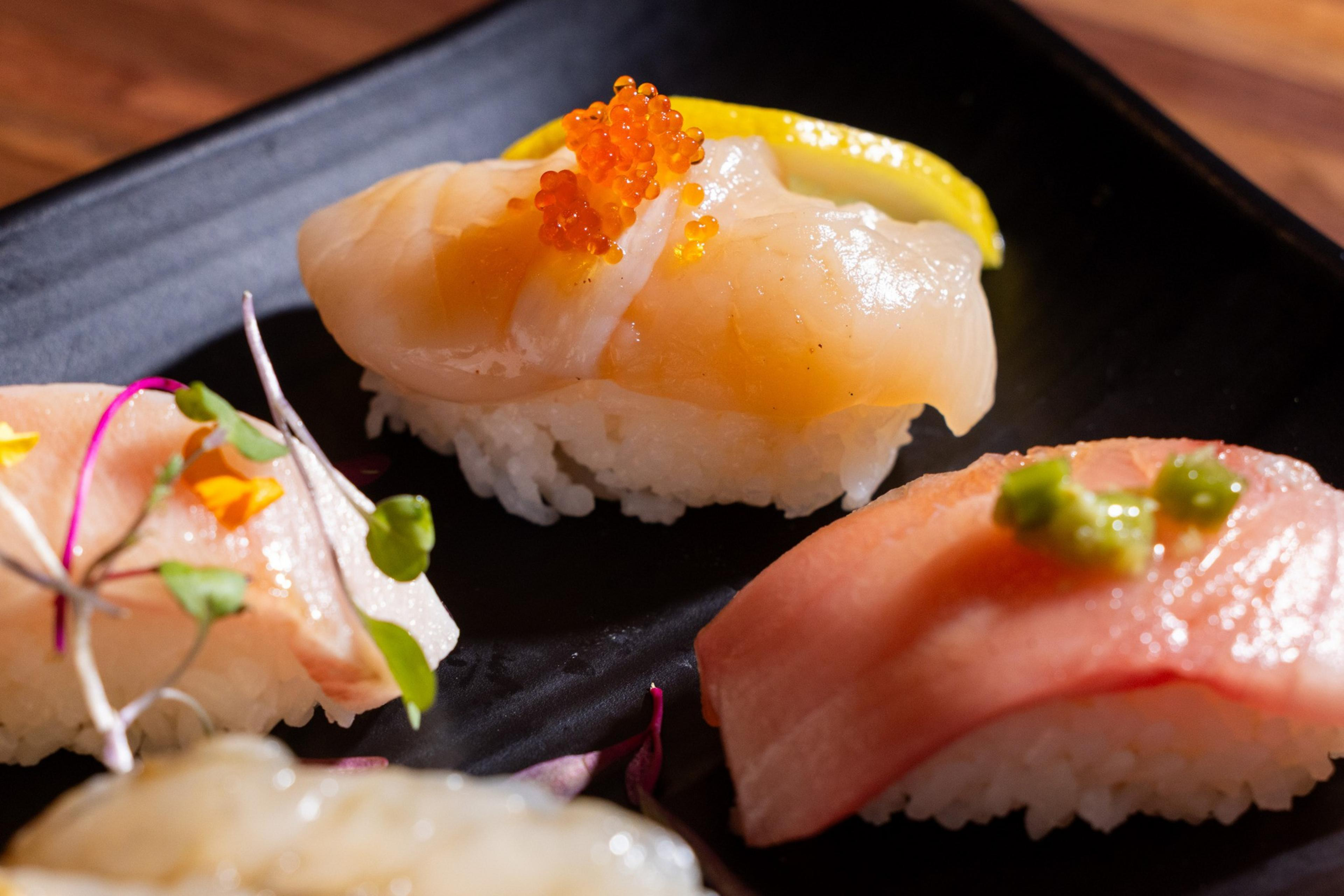 The image displays assorted sushi on a black plate. Each piece features different types of raw fish atop rice, garnished with toppings like roe and greens.