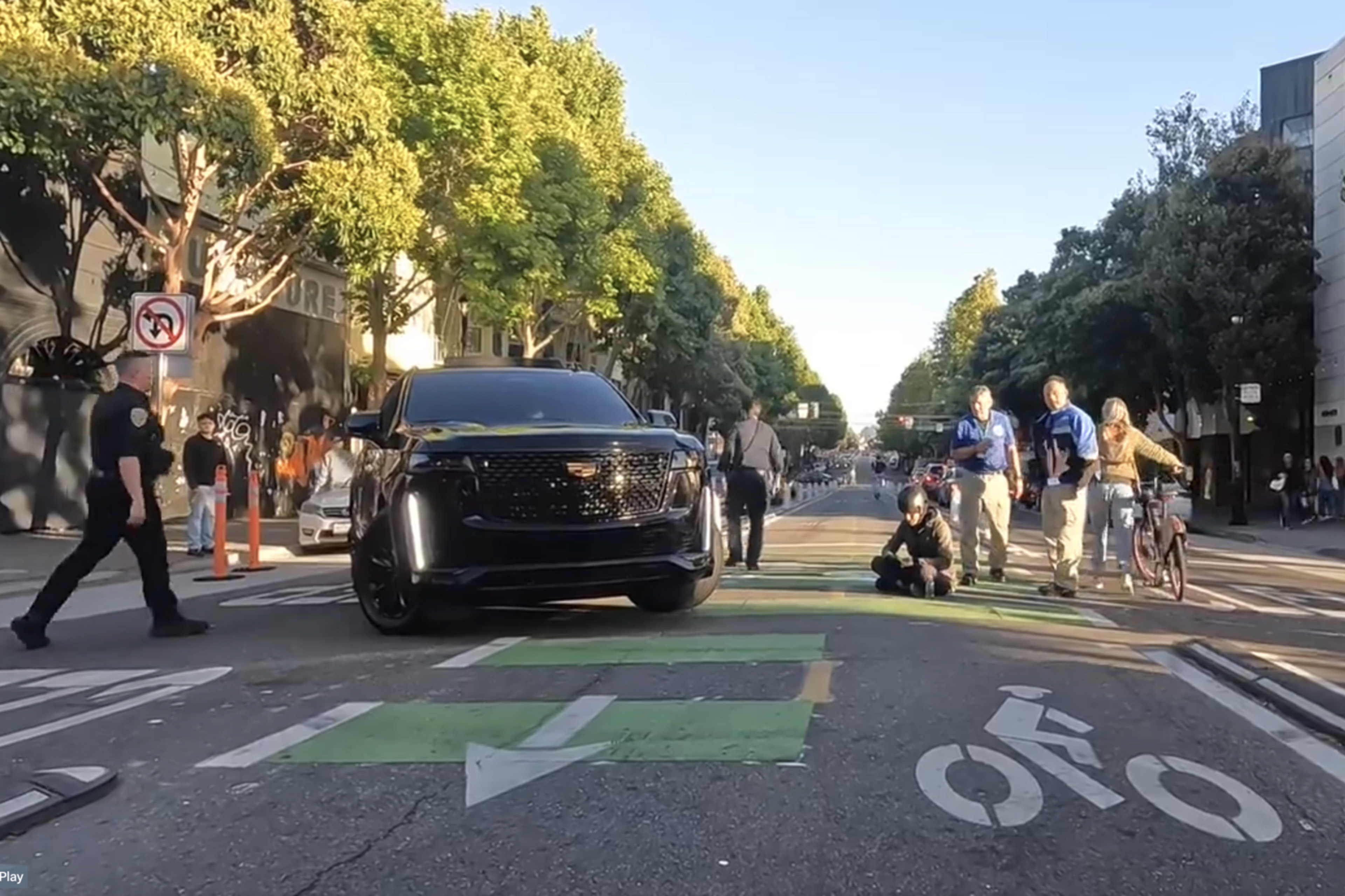 A black SUV is stopped in the middle of a city street while pedestrians and a cyclist cross. A police officer walks nearby, and the street is lined with trees and buildings.