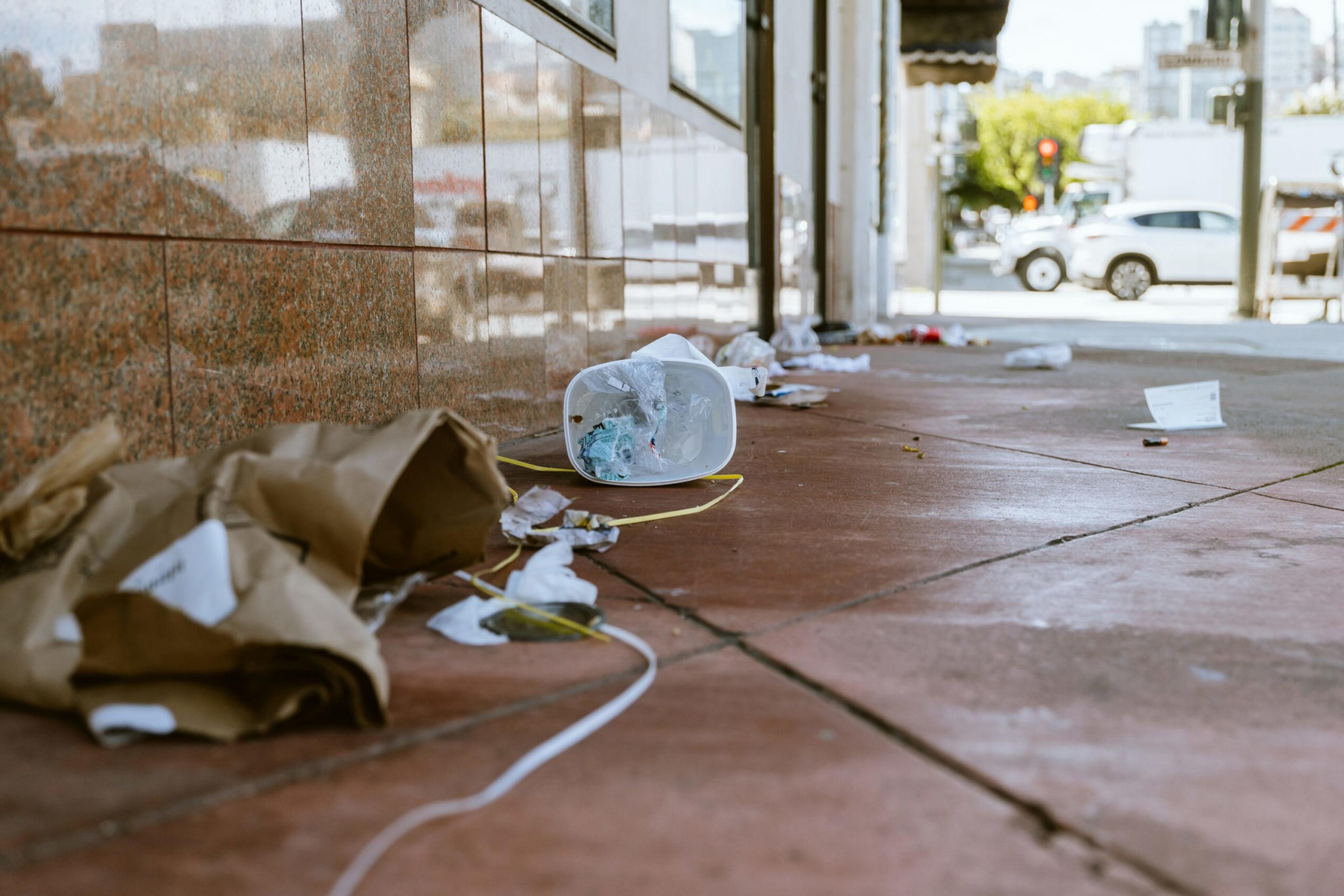 Litter lines a sidewalk: paper bags, a plastic bowl, and other trash, with a street in the background.