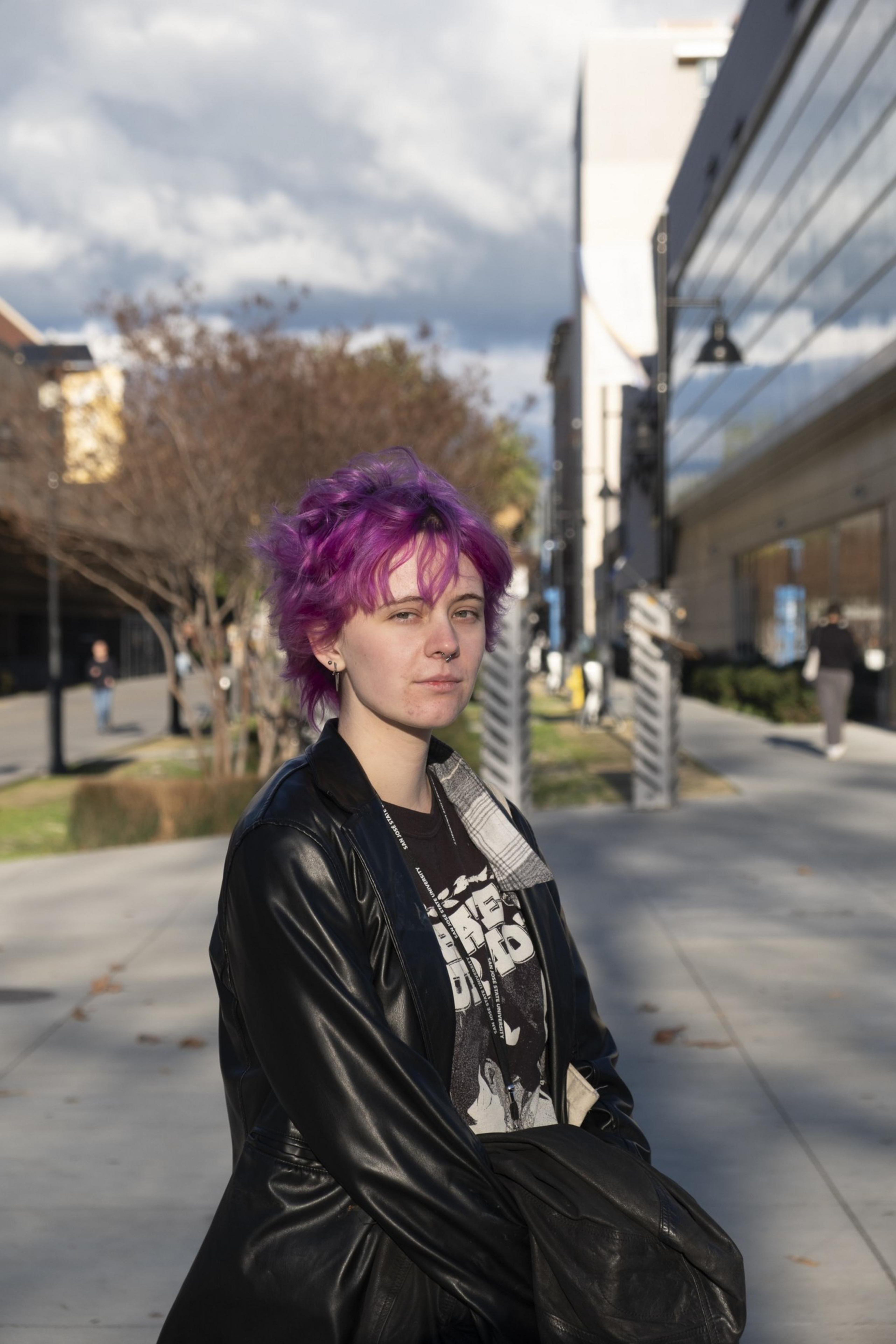 A person with bright pink hair stands on a sidewalk, wearing a black leather jacket and a graphic tee. The background includes buildings and trees under a cloudy sky.