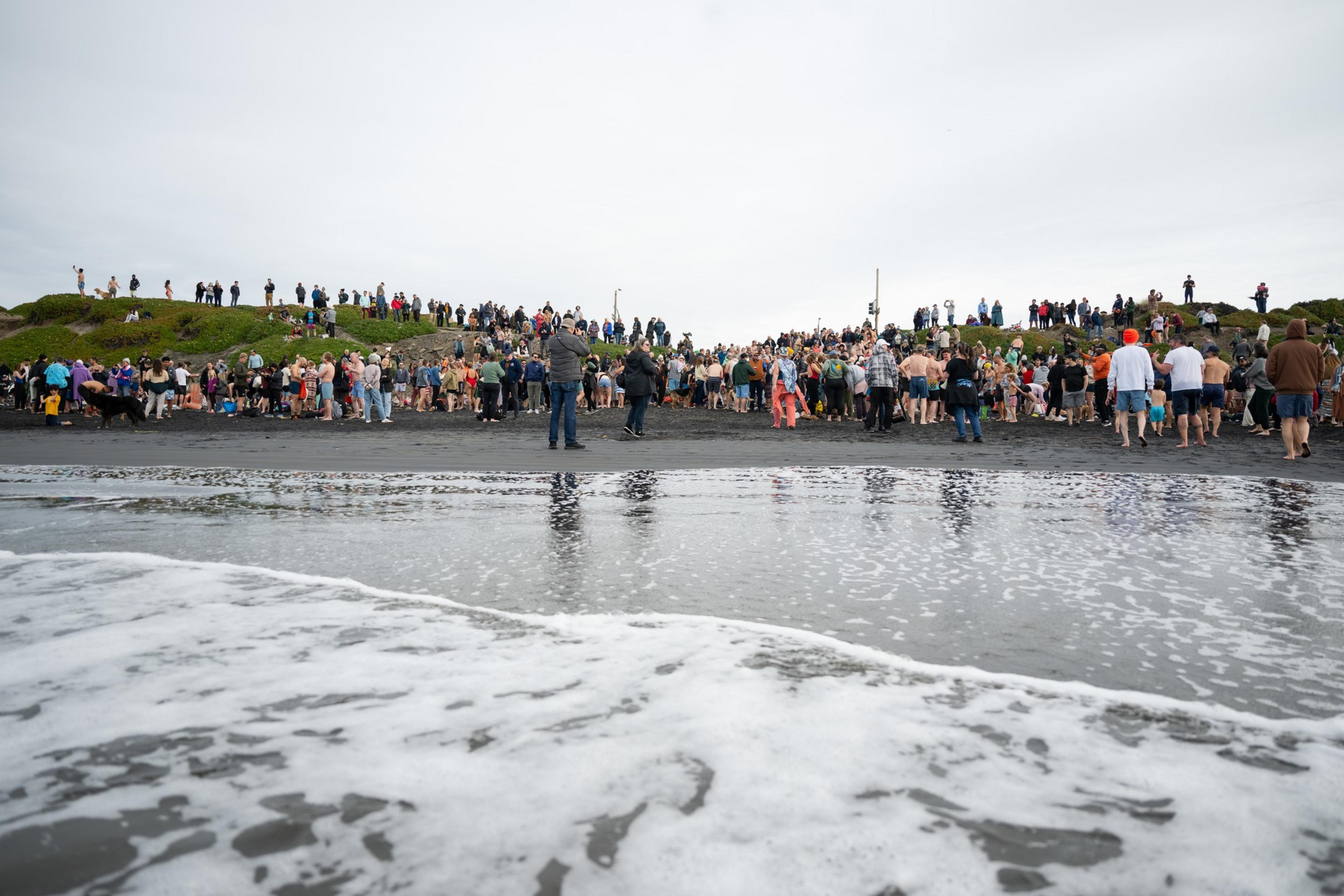 A large crowd gathers on a beach near the water's edge, with people dressed in swimsuits and casual clothes. The scene is captured from the shoreline.