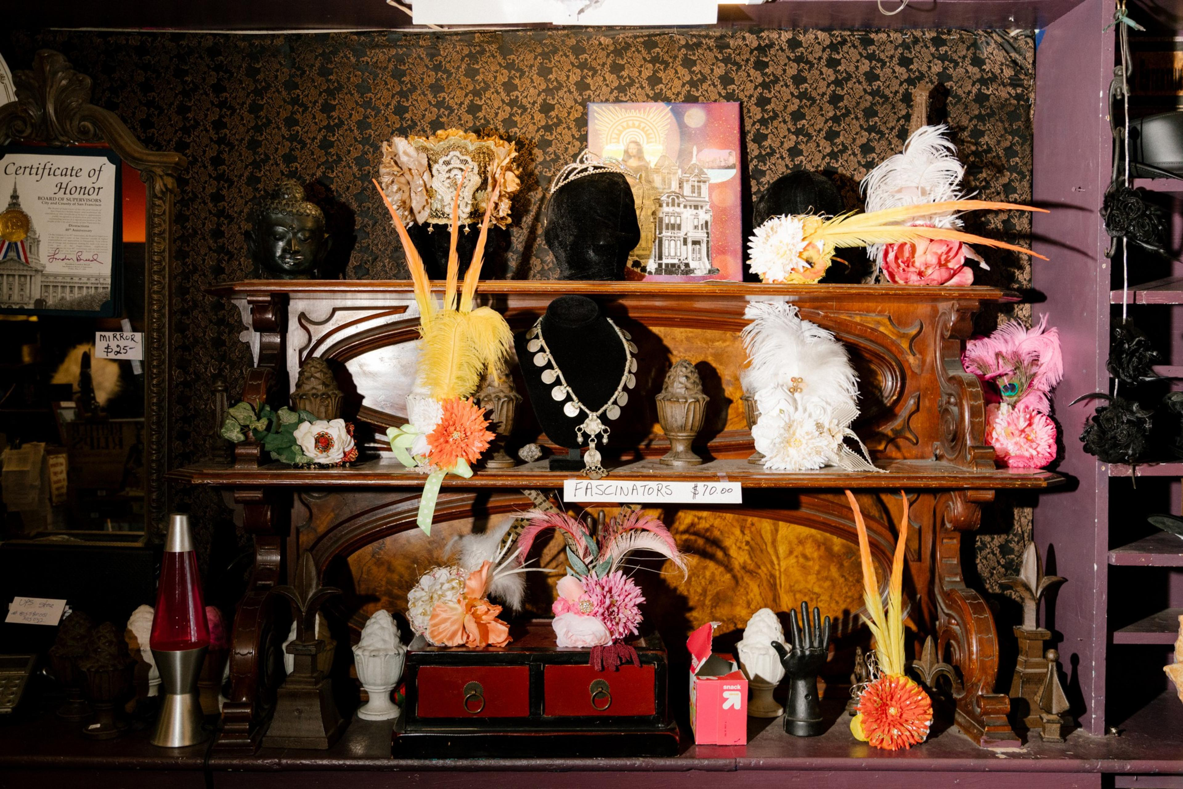 A vintage wooden shelf displays feathered fascinators, a black necklace bust with jewelry, decorative flowers, a lava lamp, and ornate small statues.