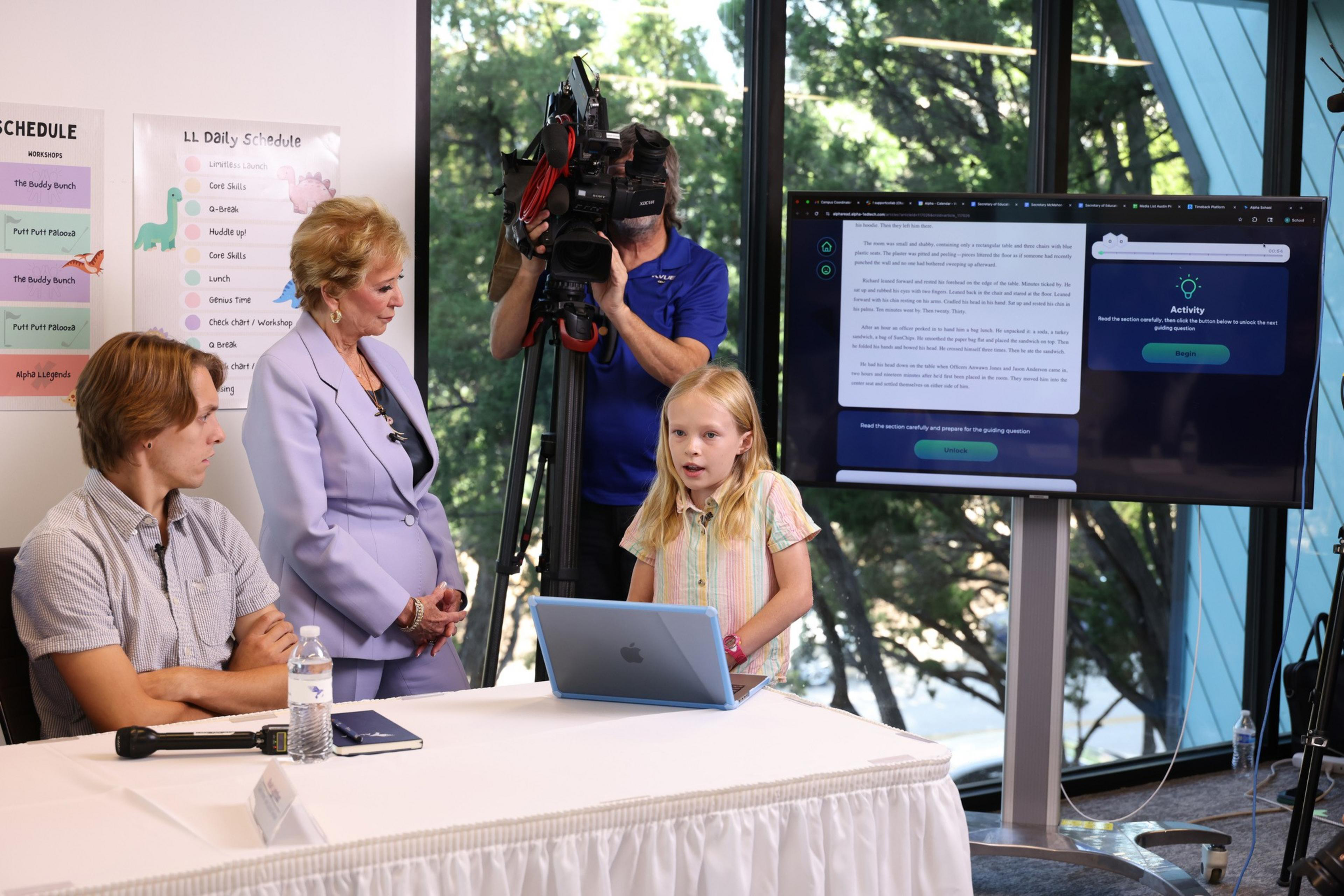 A young girl speaks at a table with a laptop while a woman, a seated boy, and a cameraman watch, with a large screen displaying text behind them.