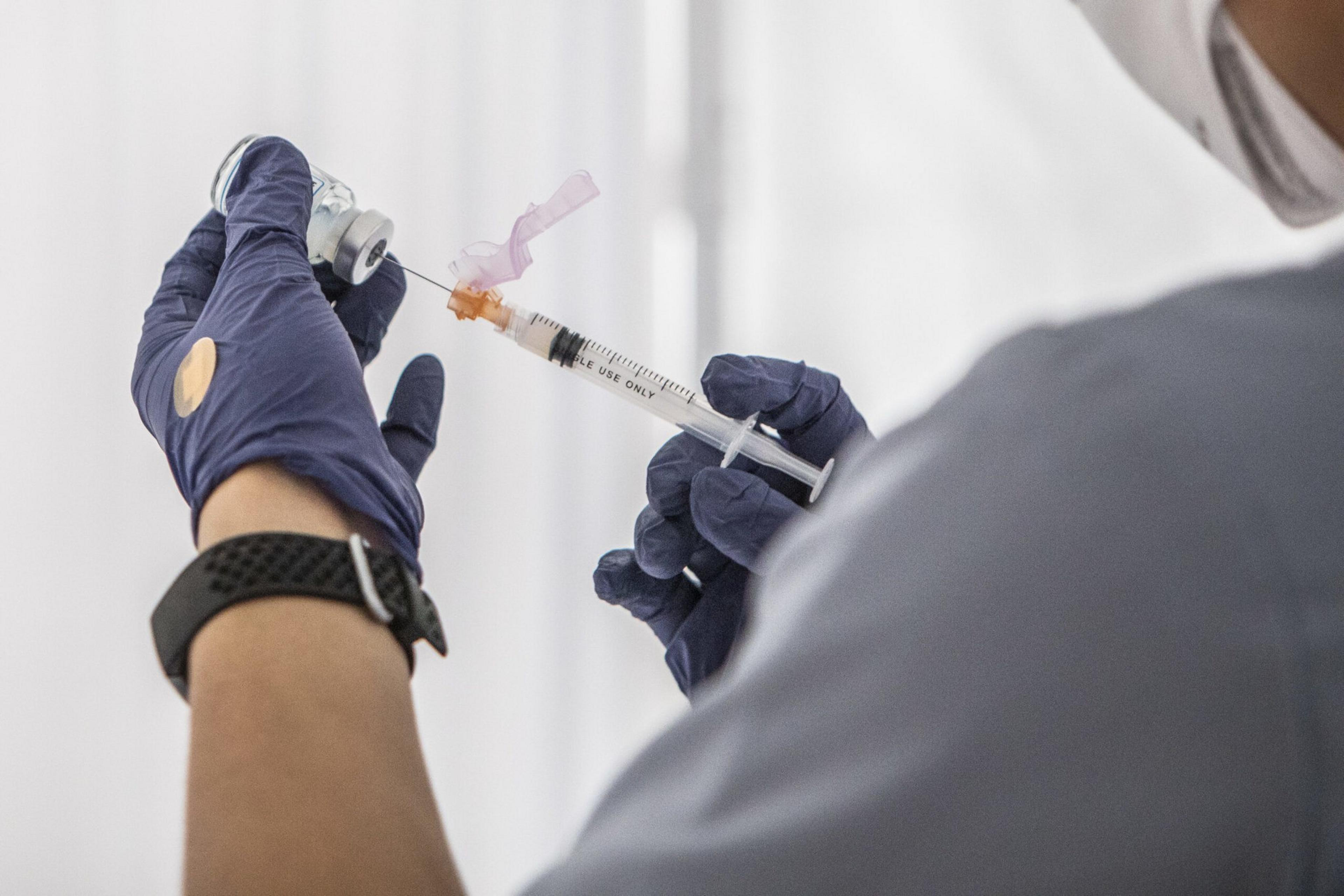 A person wearing blue gloves draws liquid from a vial into a syringe, carefully handling the needle and syringe.