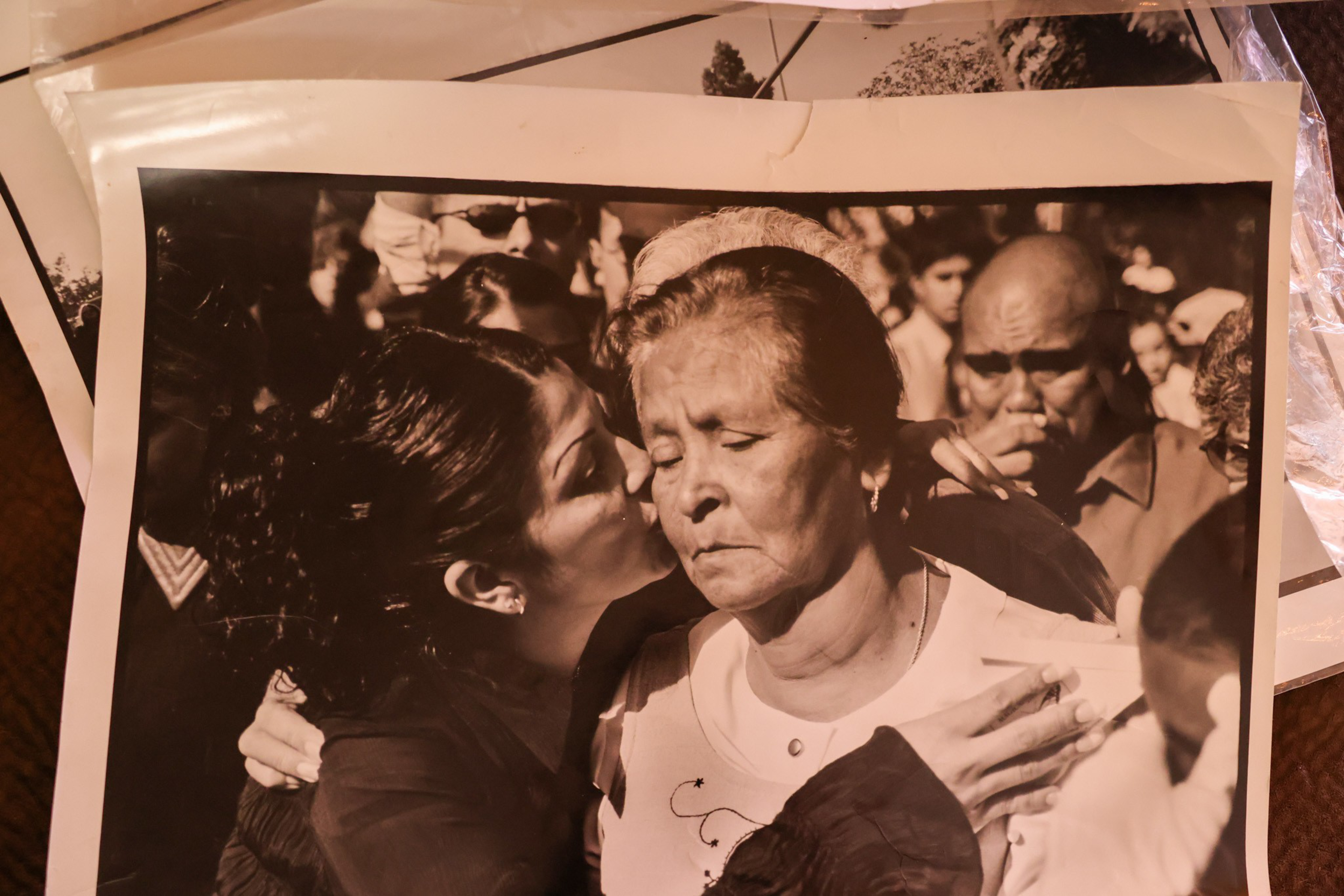 A younger woman kisses an older woman on the cheek while hugging her, surrounded by a crowd of solemn people.