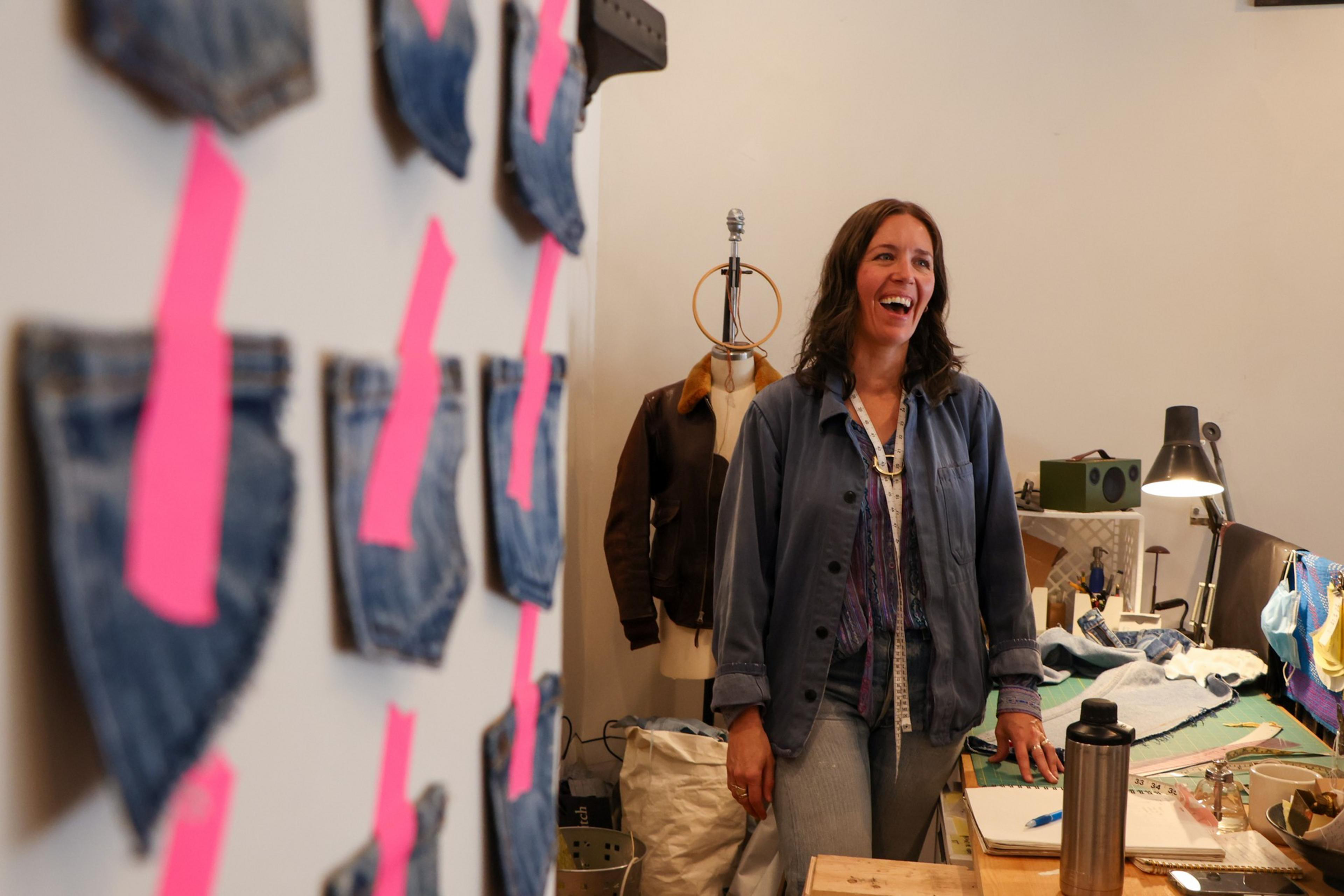 A woman in casual clothes laughs while standing in a workspace with denim fabric swatches taped to the wall and sewing supplies on the table.