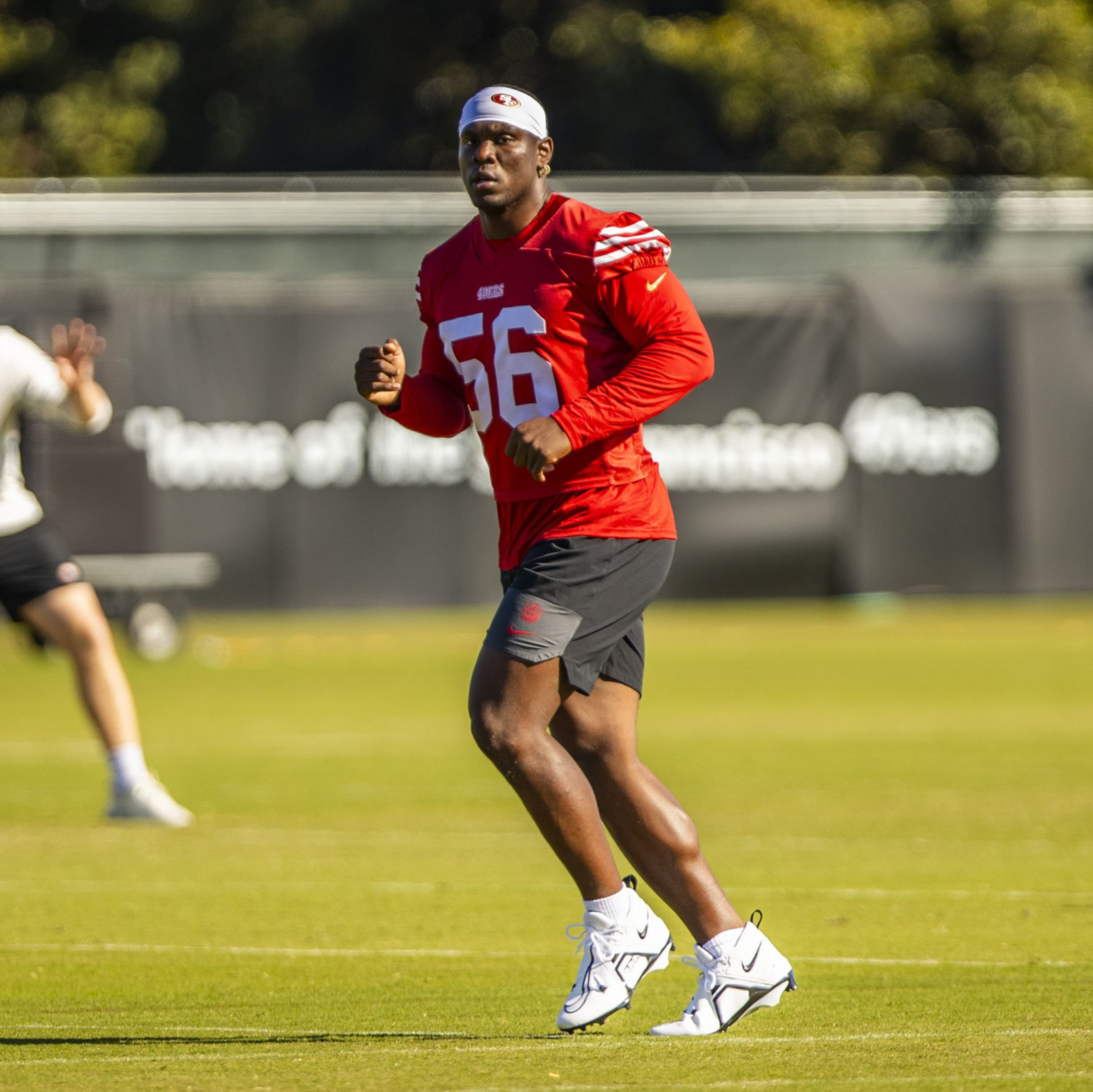A football player in a red jersey numbered 56 and black shorts runs on a green field during practice with trees and a banner in the background.