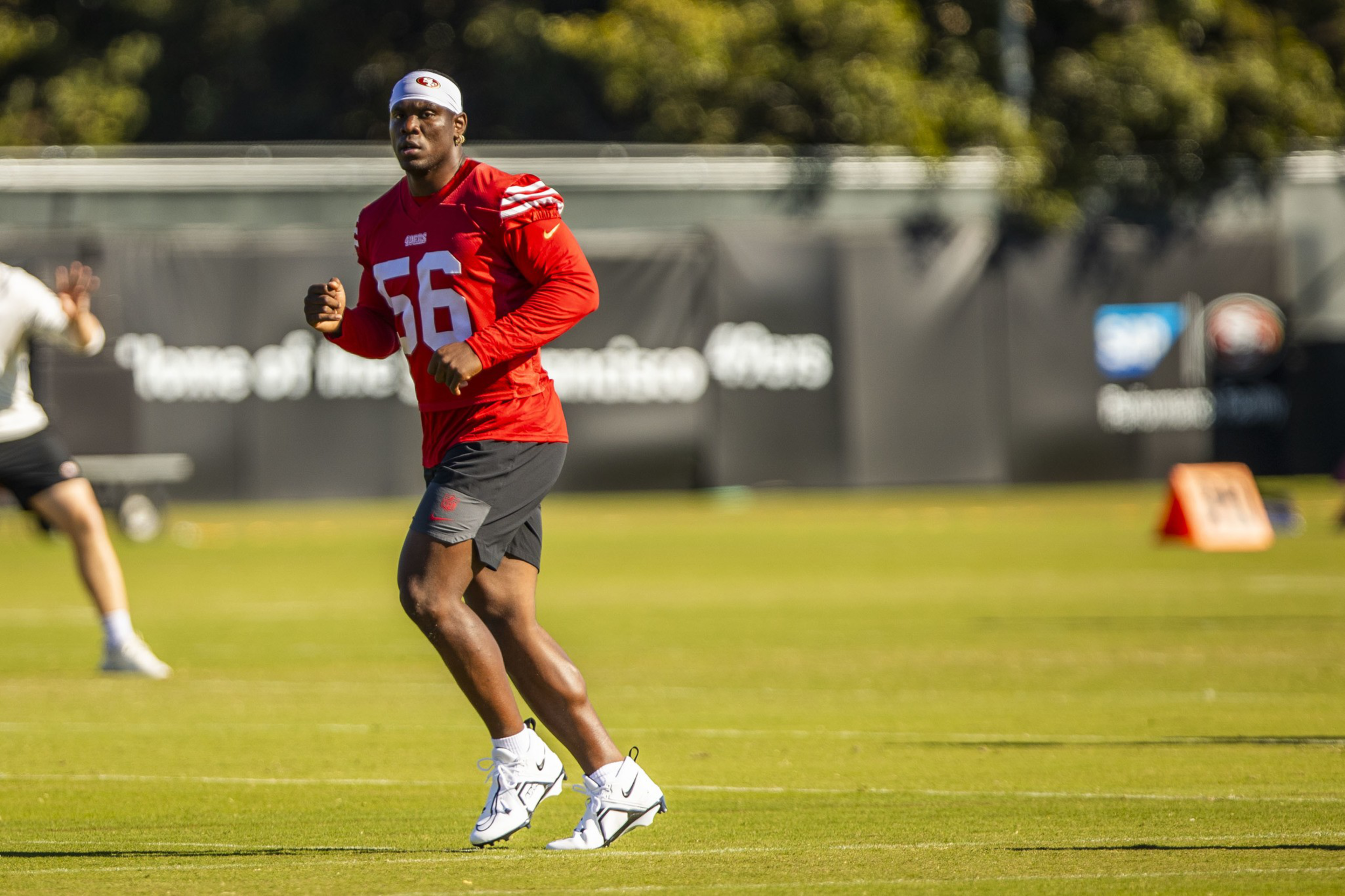 A football player in a red jersey numbered 56 and black shorts runs on a green field during practice with trees and a banner in the background.