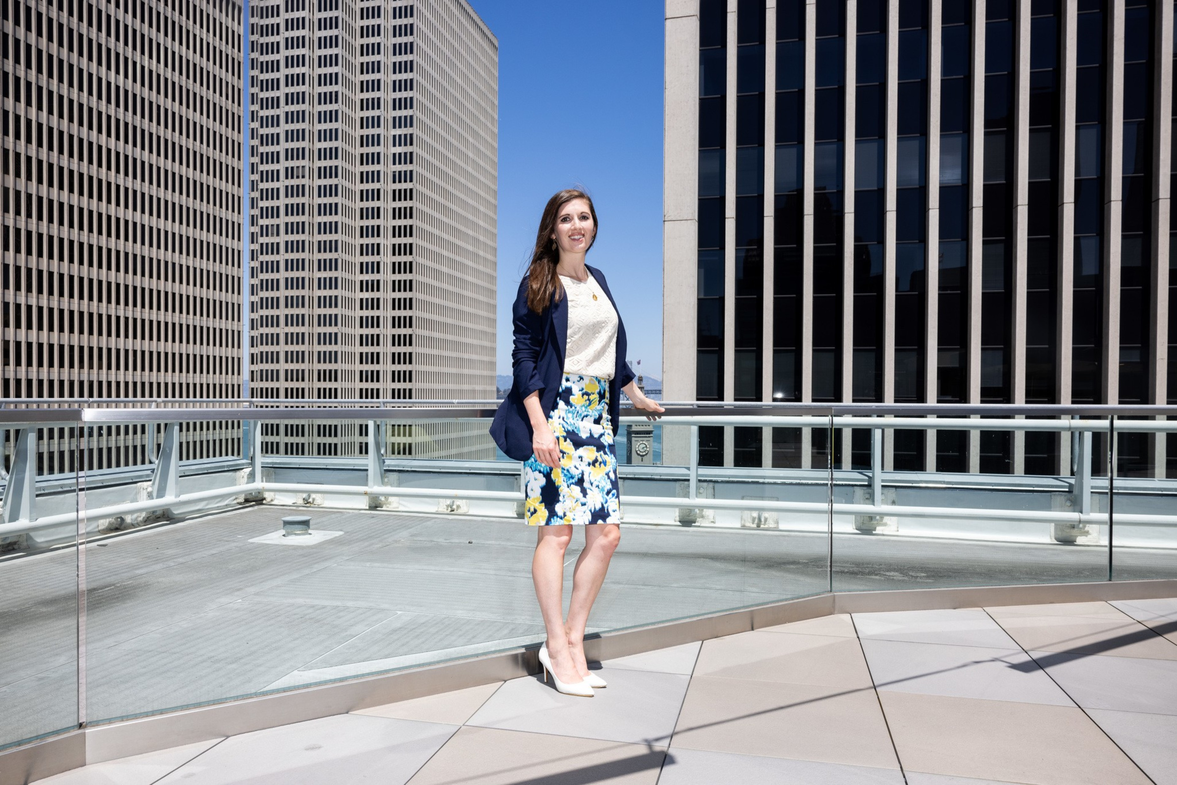 A woman stands on a rooftop, surrounded by tall buildings. She is dressed in a navy blazer, white blouse, a floral skirt, and white heels, smiling at the camera.