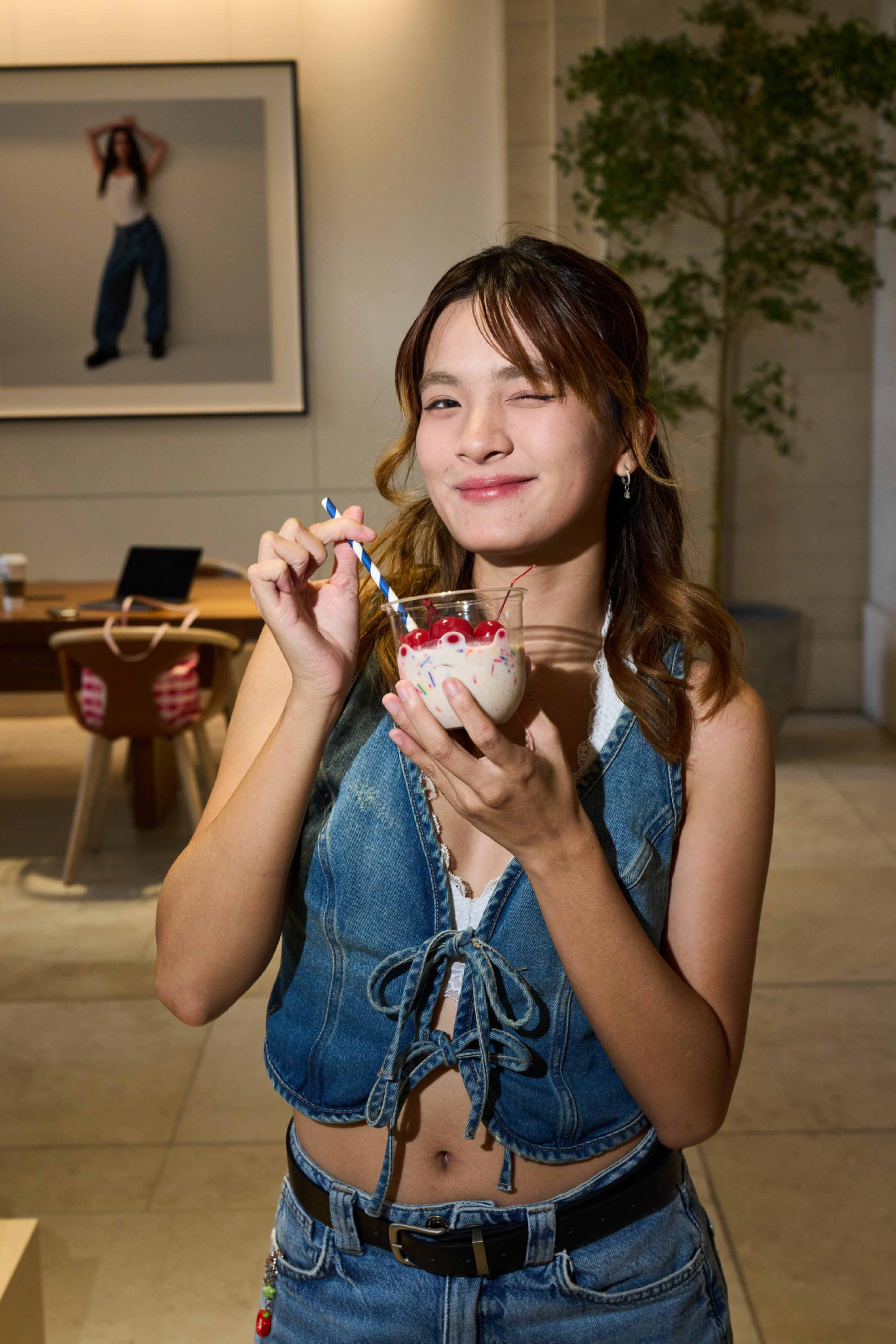 A young woman winks and smiles while holding a dessert topped with cherries and stirring it with a striped blue and white spoon.