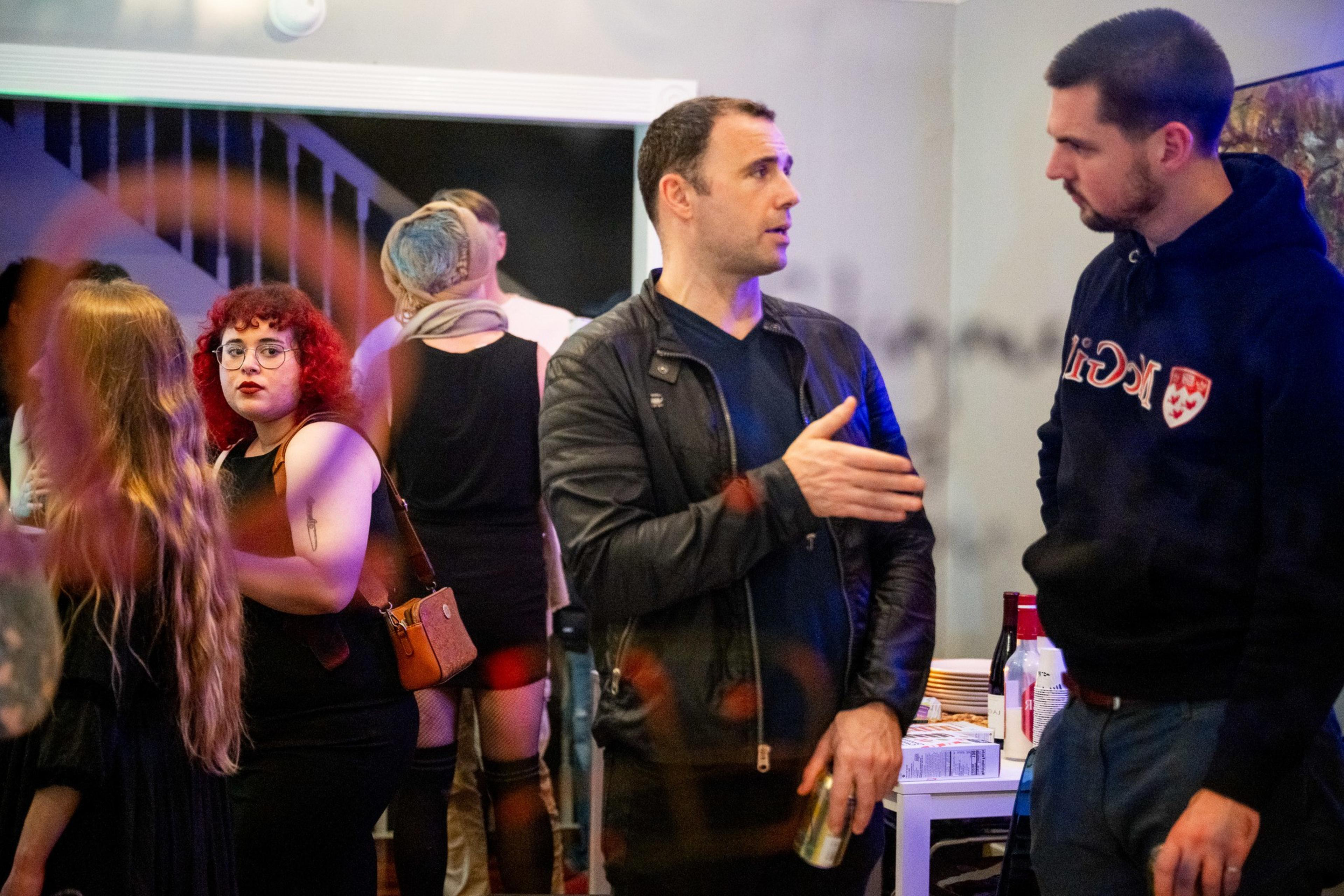 People are gathered indoors at a social event. A woman with curly red hair looks towards the camera, while two men converse nearby. Drinks are on a table.