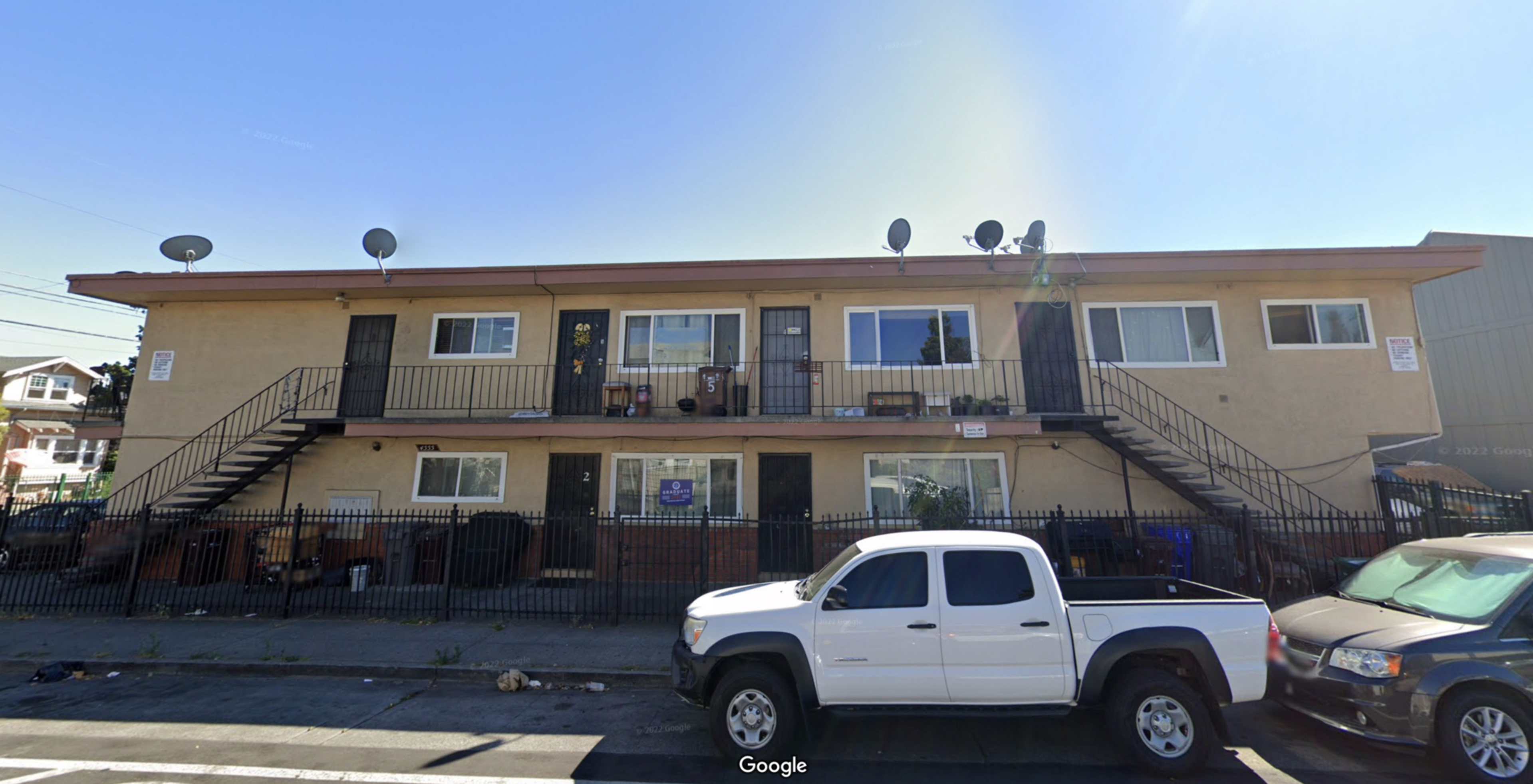 A two-story beige apartment building with external black metal staircases, iron railings, satellite dishes, and cars parked in front on a sunny day.