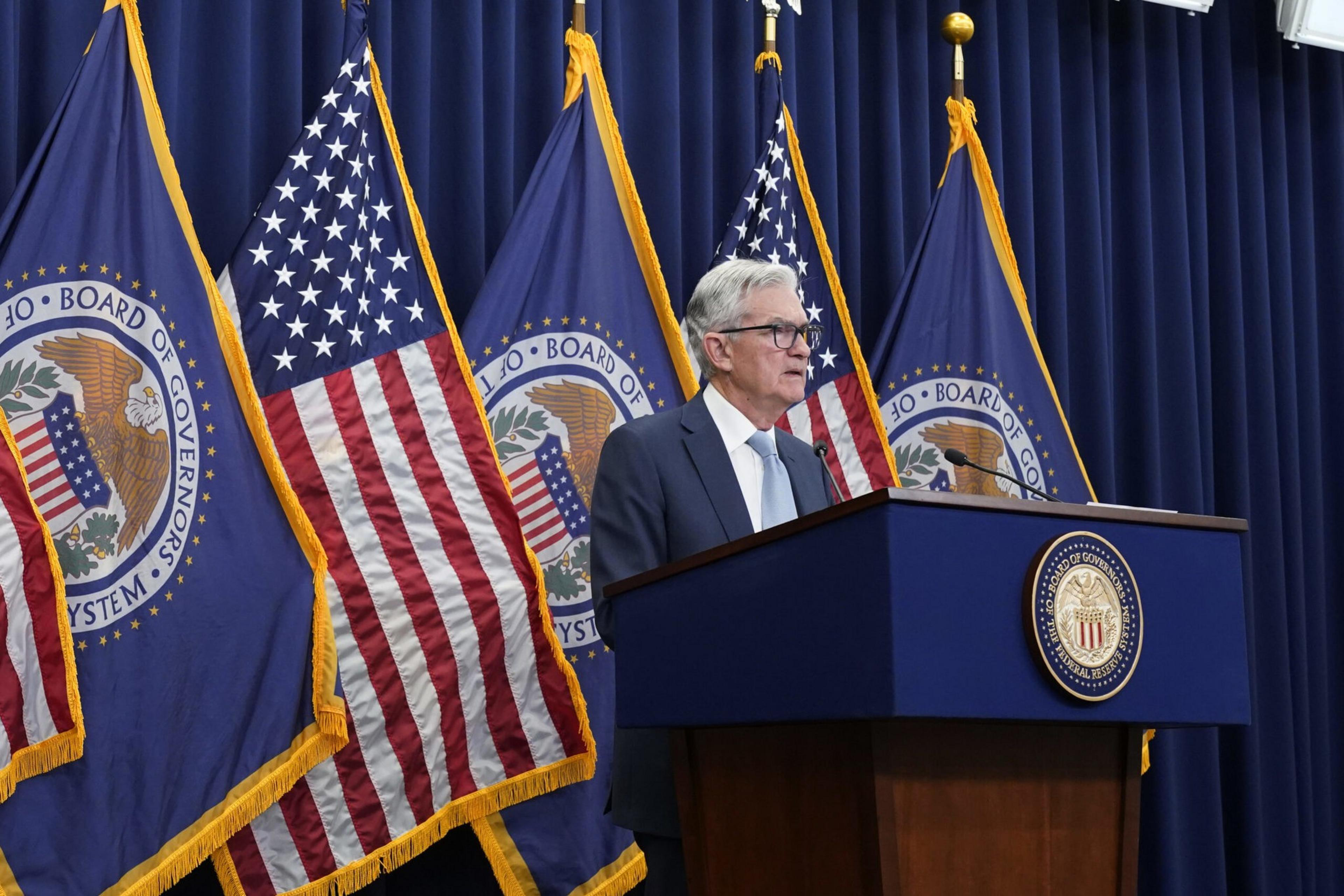 An older man in a suit stands at a podium with the Federal Reserve's seal, flanked by U.S. and Federal Reserve flags, speaking against a backdrop of blue curtains.
