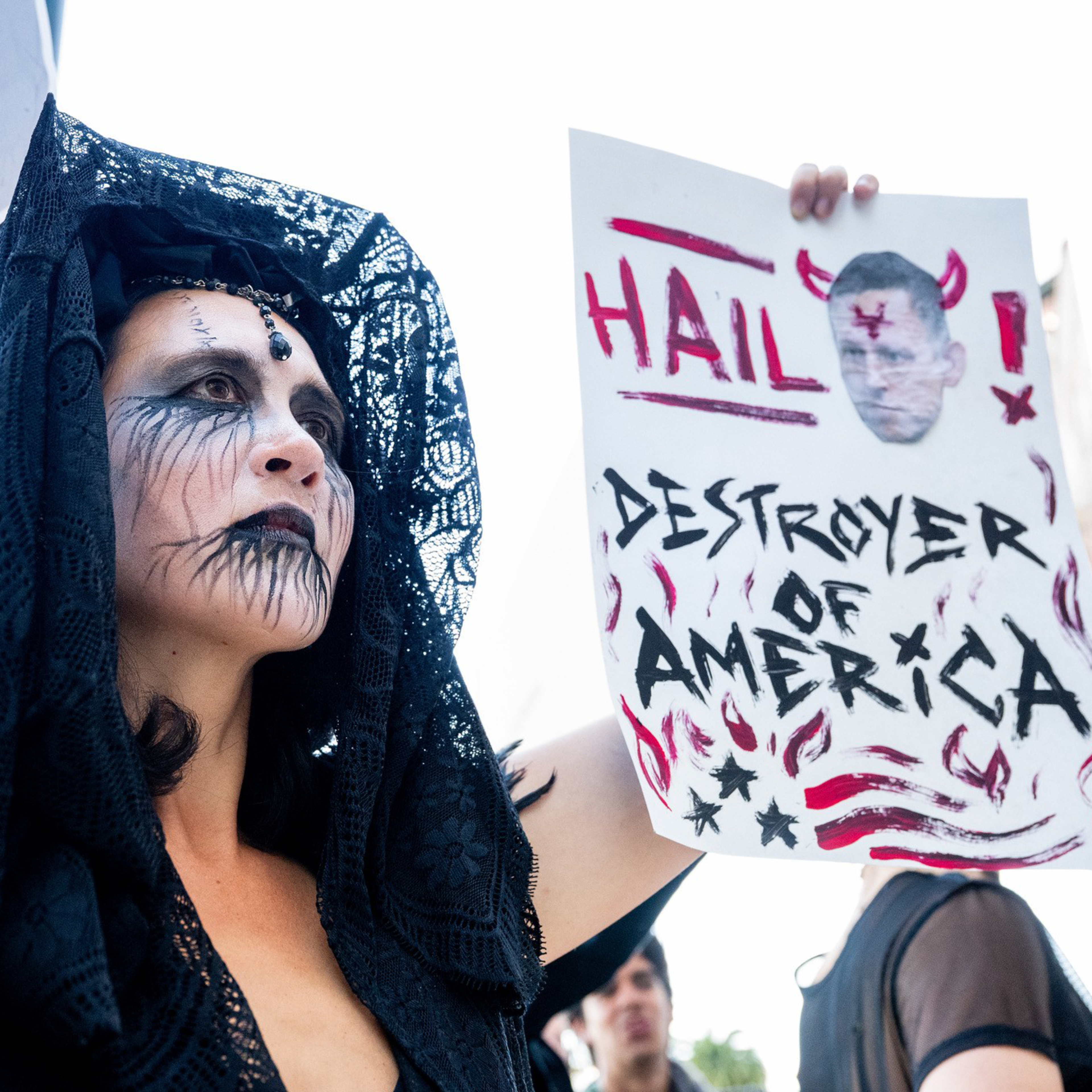 A person with black lace veil and dark, smeared face paint holds a sign that reads "HAIL! DESTROYER OF AMERICA" with a photo of a man with devil horns.