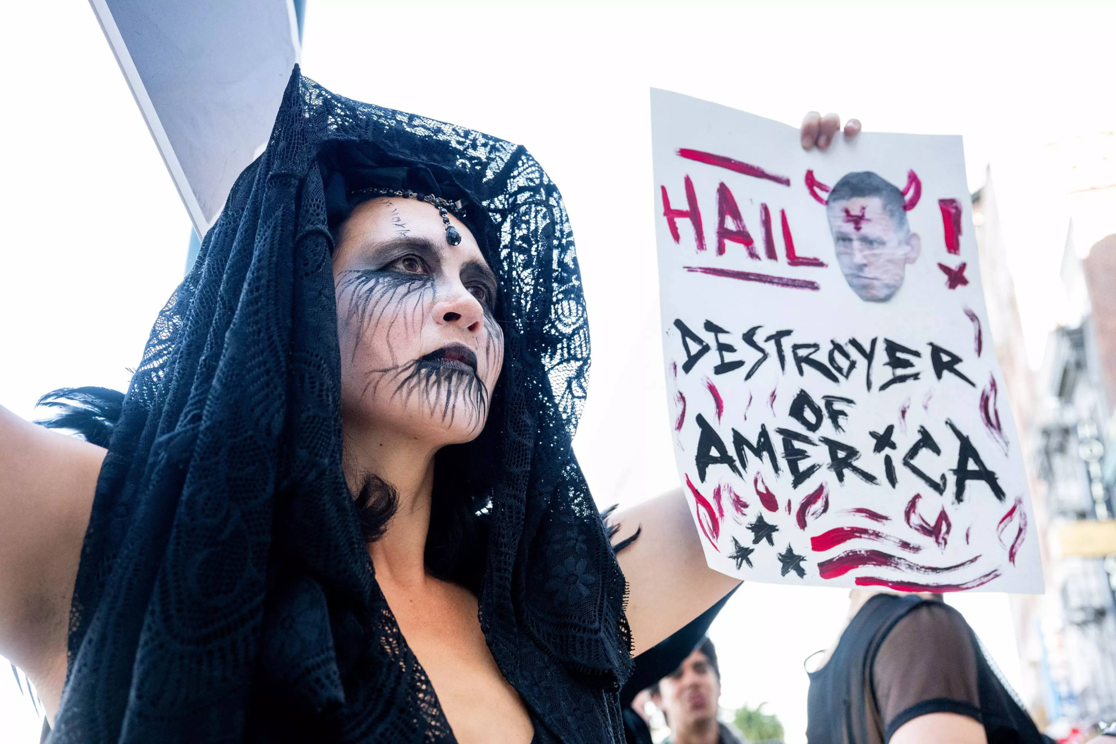 A person with black lace veil and dark, smeared face paint holds a sign that reads “HAIL! DESTROYER OF AMERICA” with a photo of a man with devil horns.