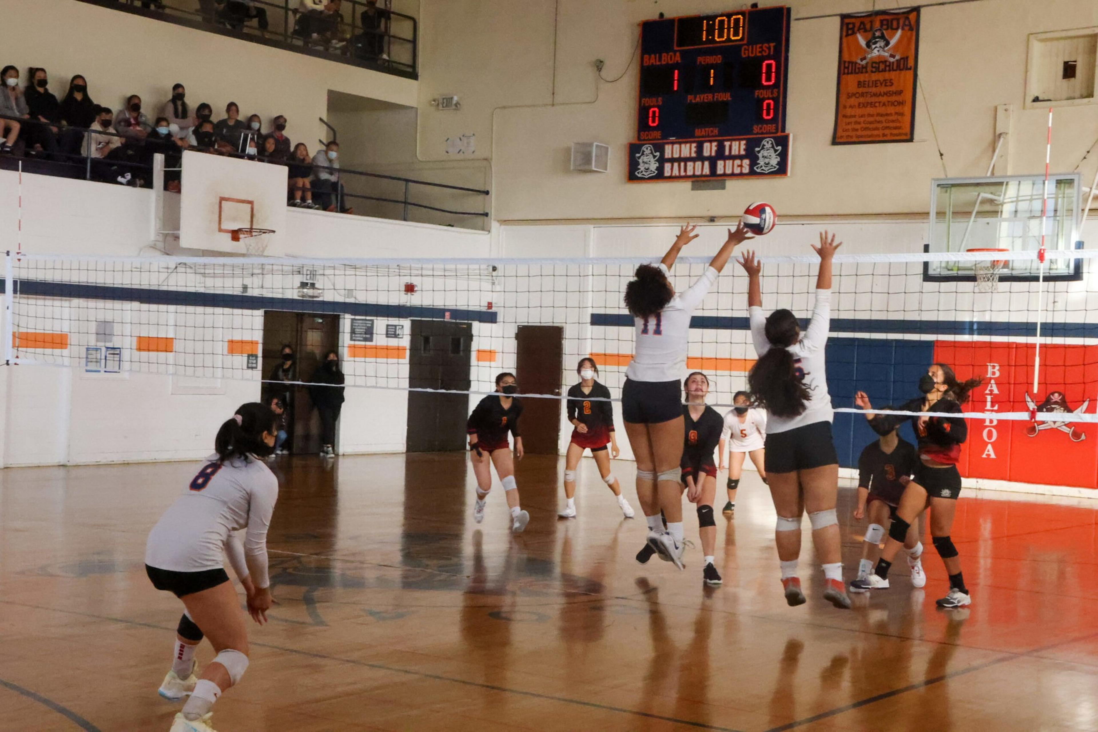 Two volleyball players in white jerseys jump to block a ball while teammates and opponents watch on a gym court with spectators in the bleachers.