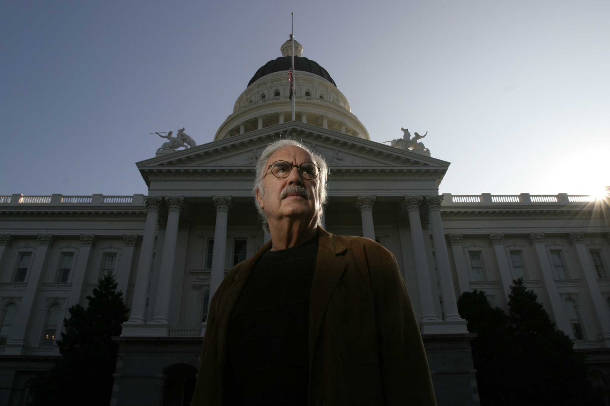 An older man with glasses and a mustache stands in front of a large domed government building during sunset.