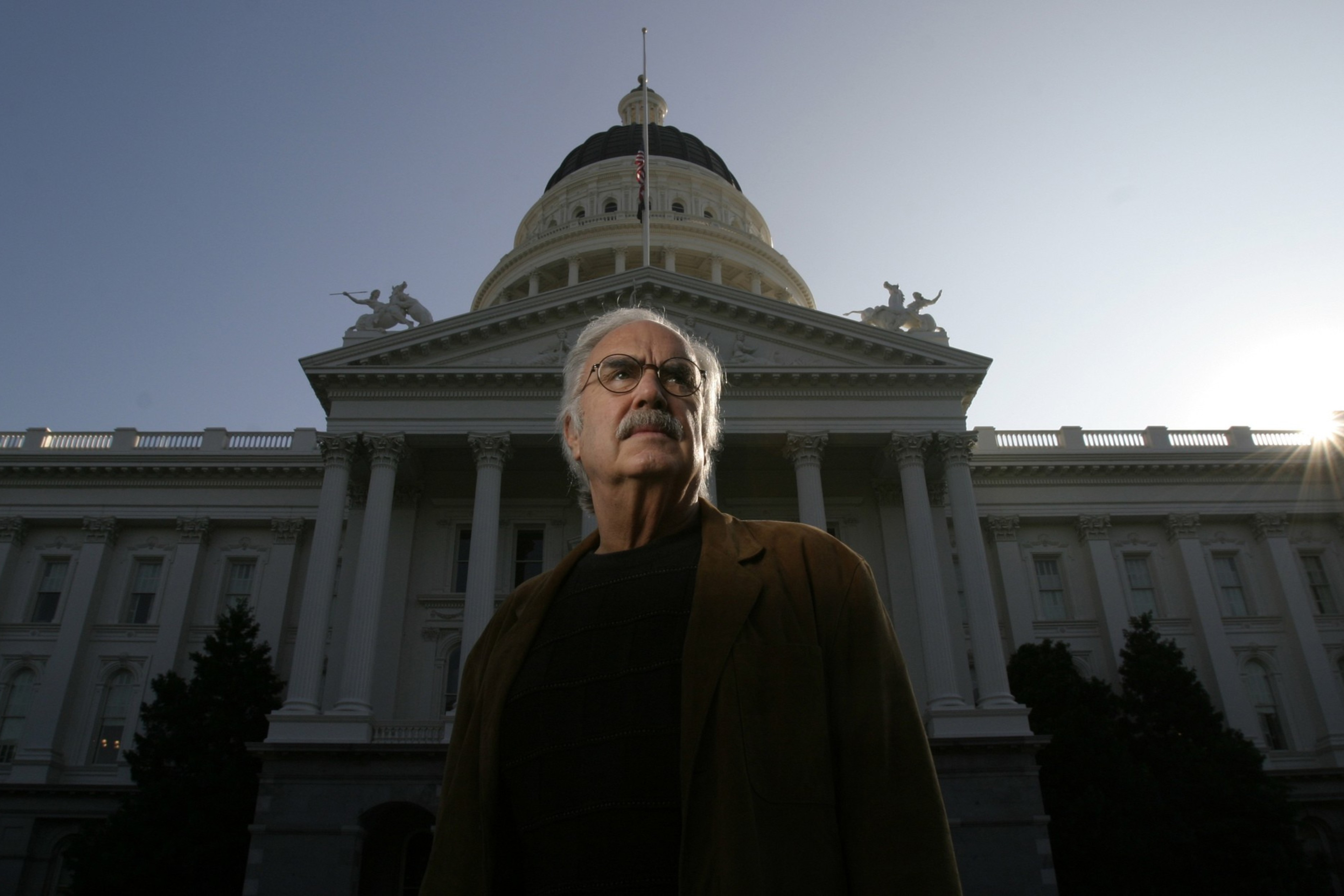 An older man with glasses and a mustache stands in front of a large domed government building during sunset.