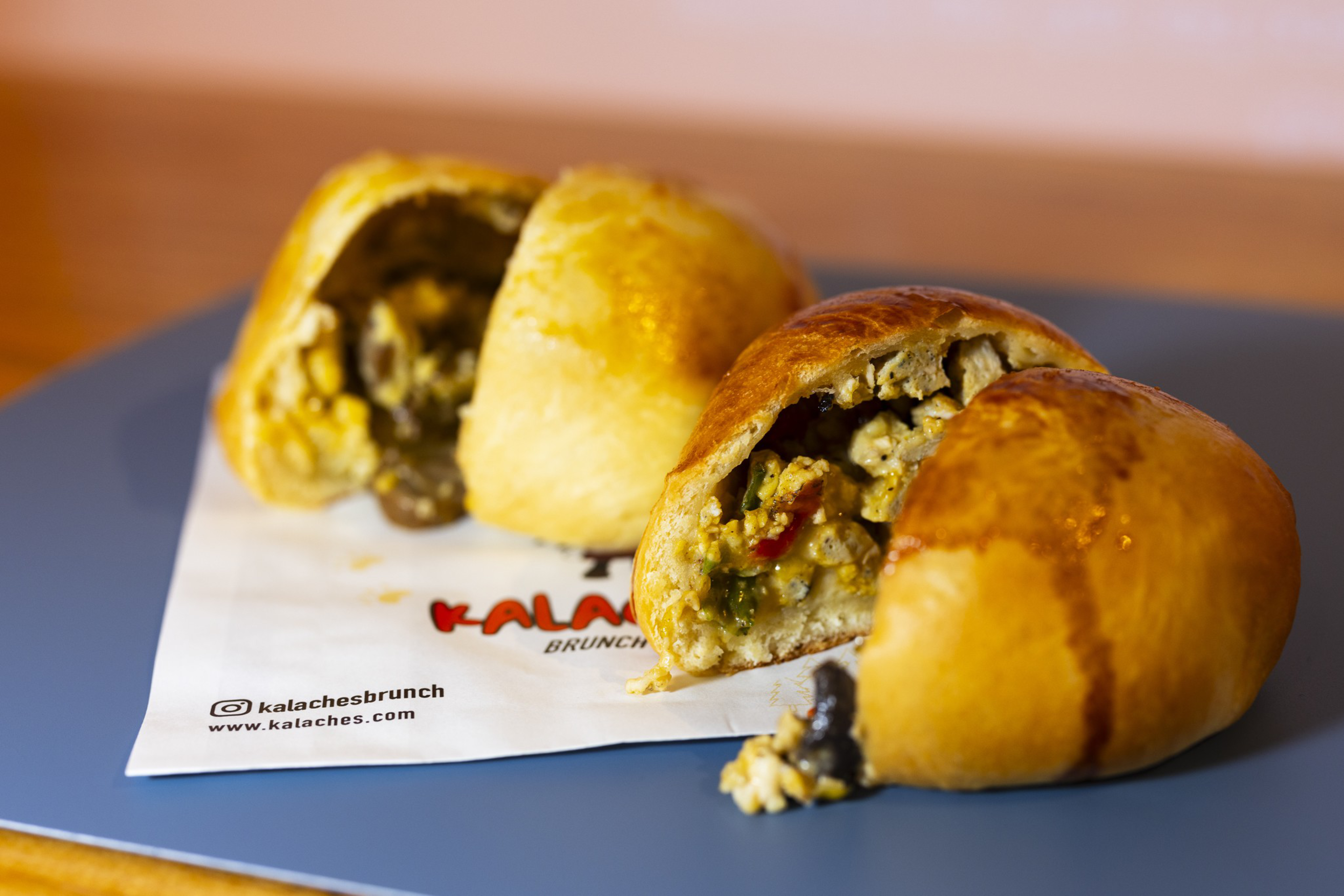 Two golden-brown kalaches are cut open, revealing a savory filling of scrambled eggs, vegetables, and sausage on a paper with branding.