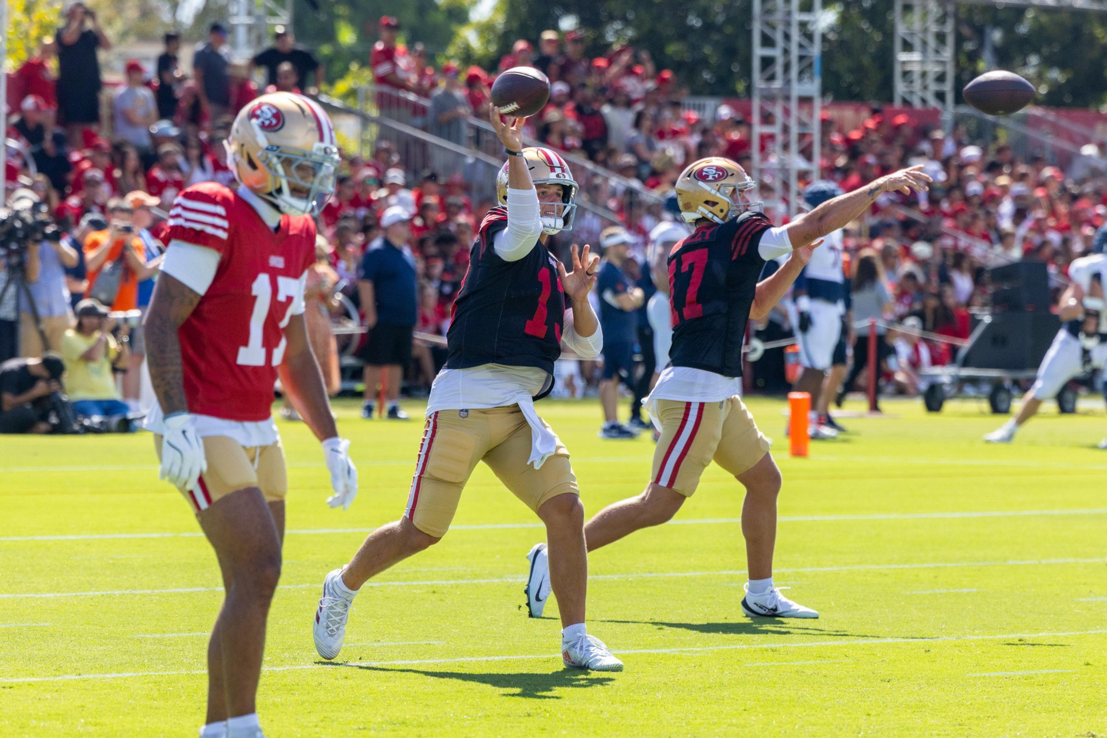 Two football players in black jerseys and gold pants are throwing footballs during practice, while another player in a red jersey stands nearby.