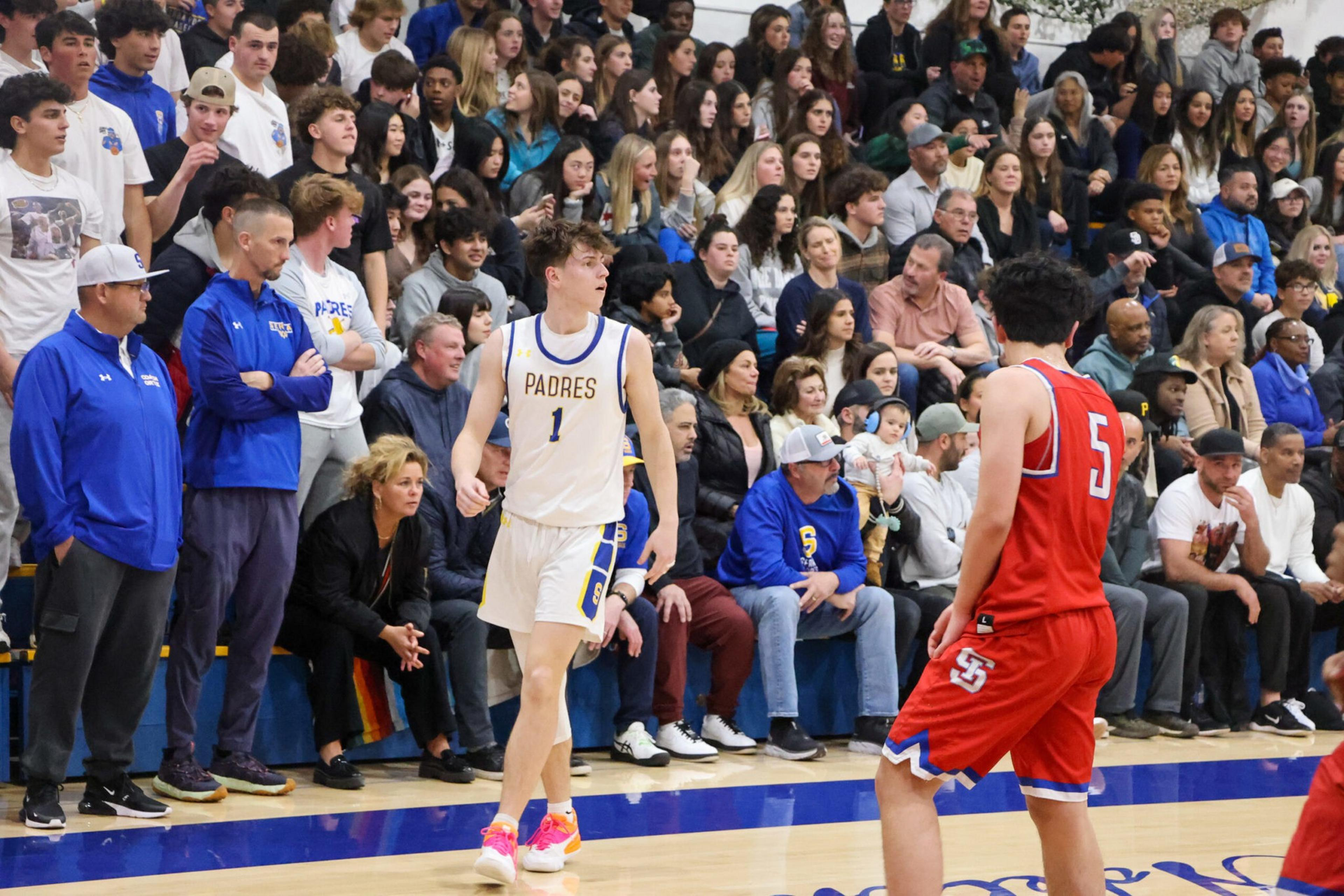 A basketball player in a white Padres jersey faces an opponent in a red jersey with number 5, as a large crowd watches from the bleachers.
