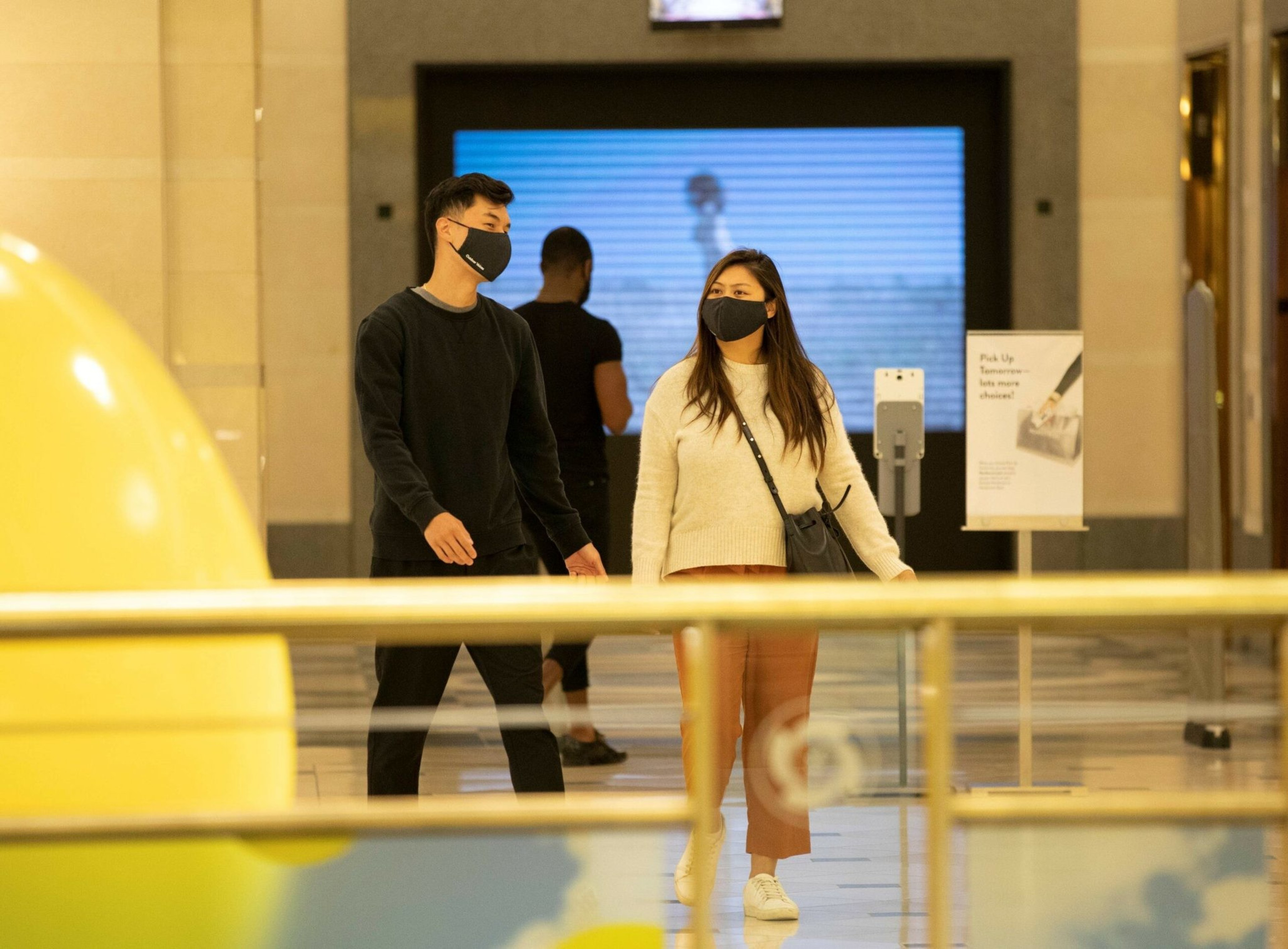A man and woman wearing black face masks walk indoors near a railing, with another person and a sign visible in the background.