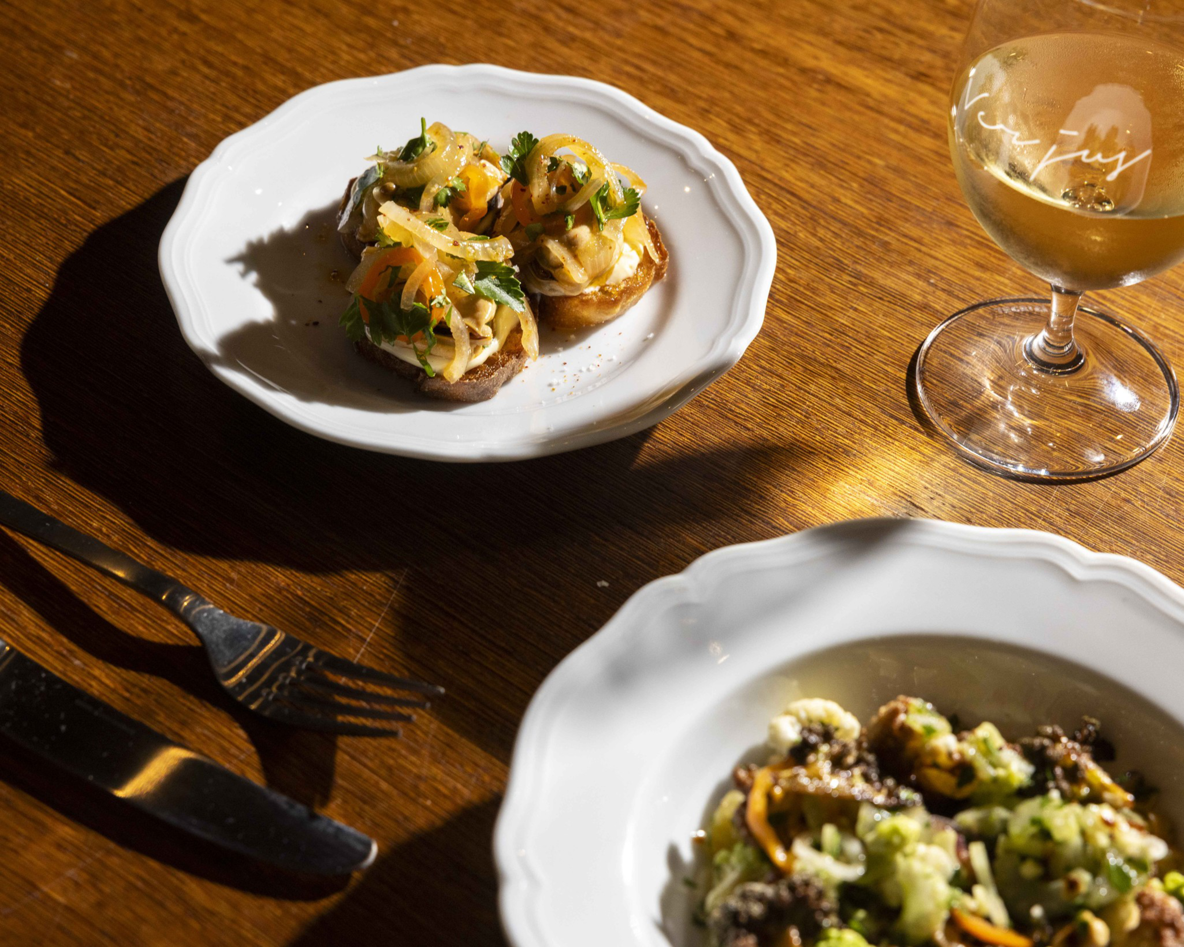 The image shows two white plates with appetizers, a glass of white wine, and silverware on a wooden table, all bathed in warm light.