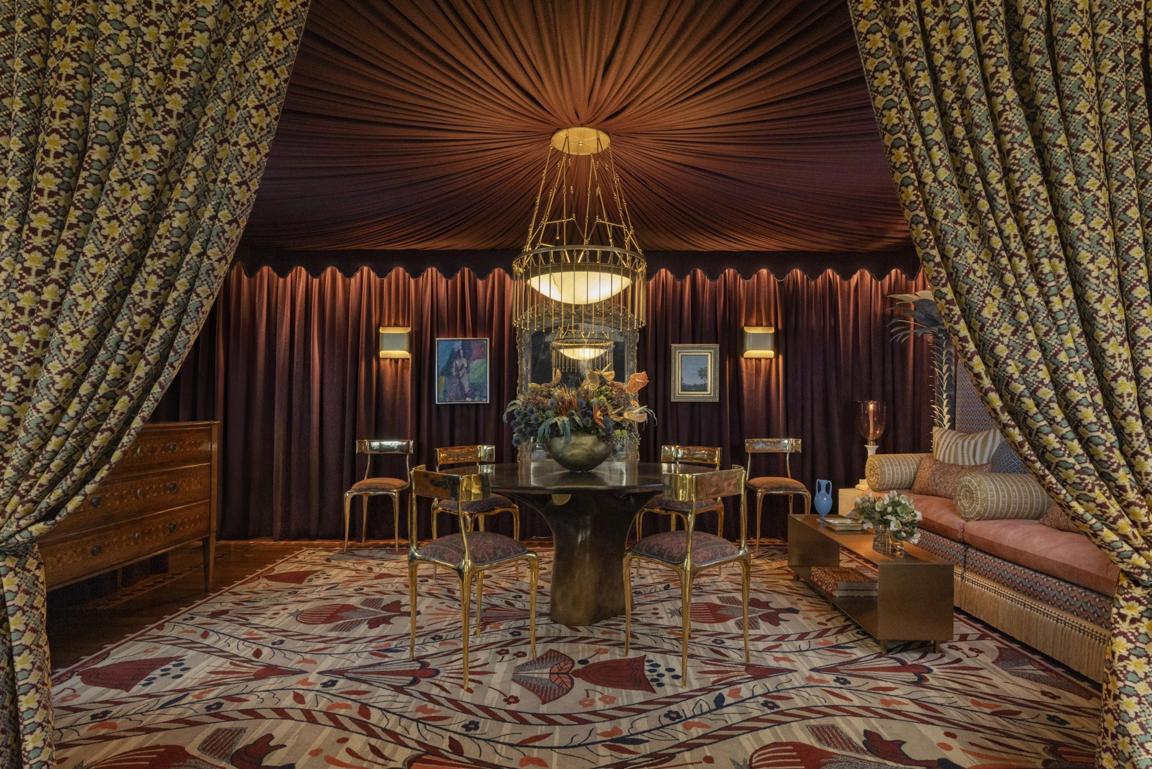 A cozy dining room features gold-accented chairs around a dark round table, patterned curtains, a large floral centerpiece, and warm orange and brown tones.
