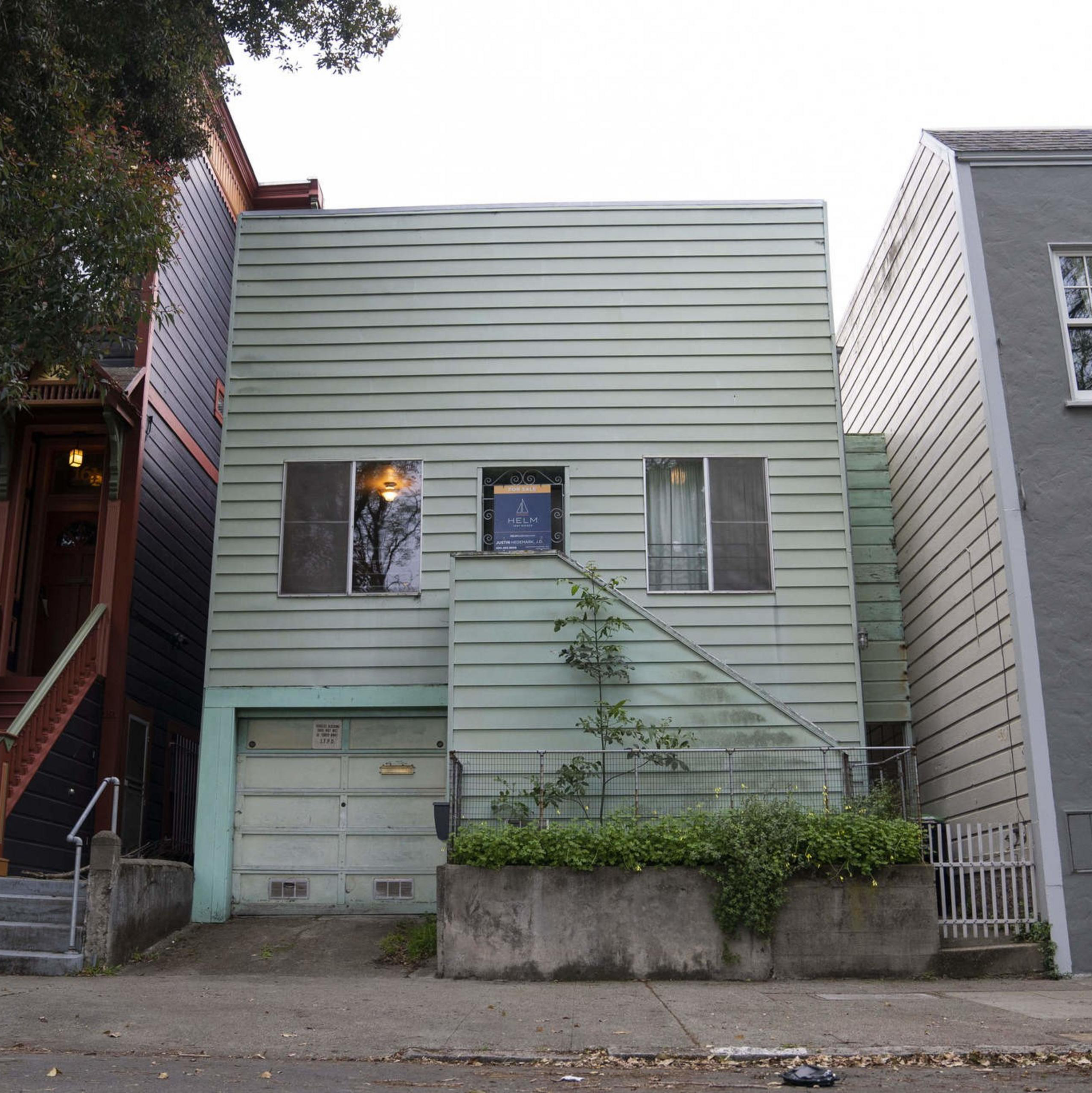 A small light green house with horizontal siding stands between two larger homes. It has a garage, two windows, and a simple staircase leading to the front door.
