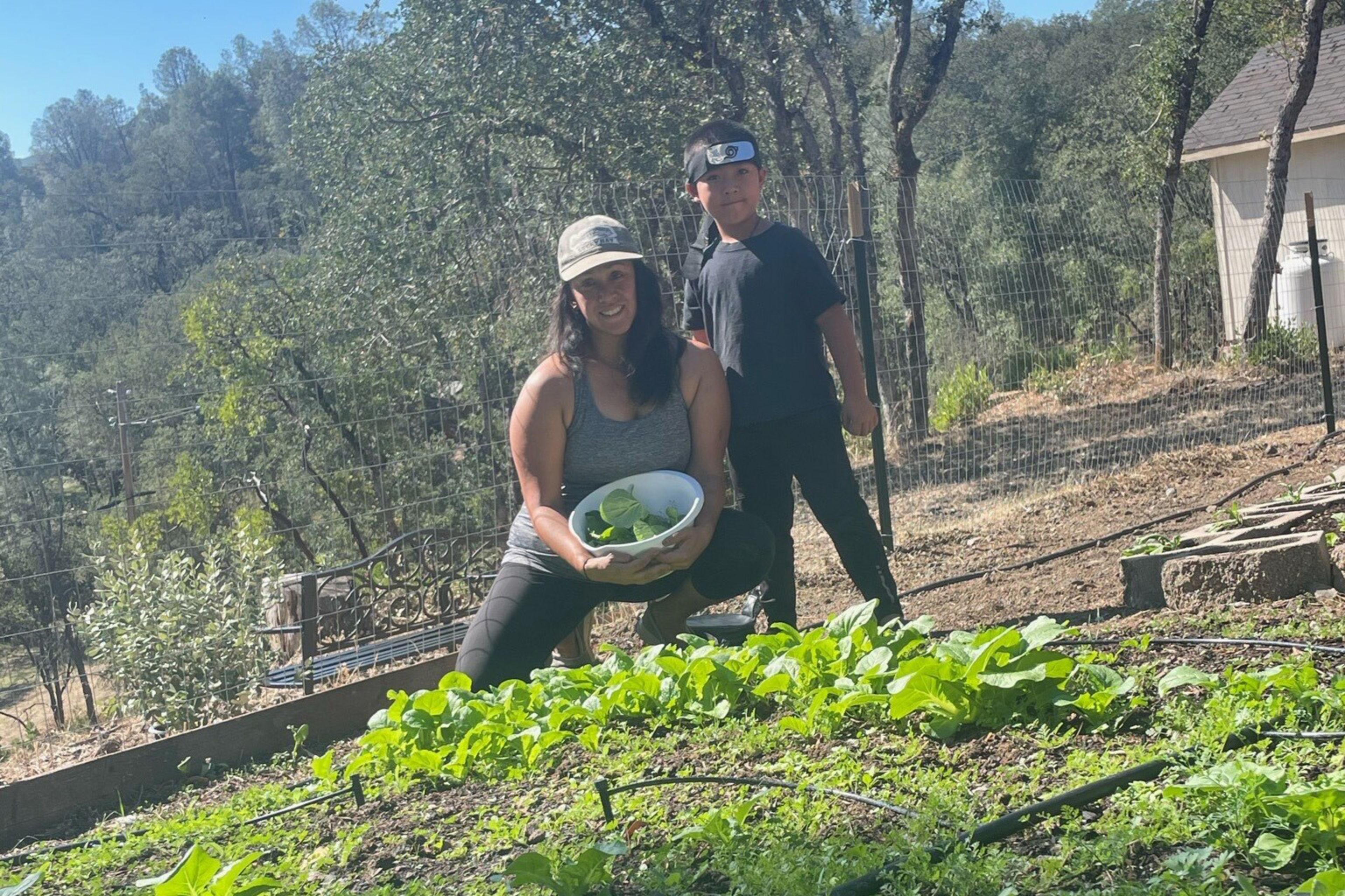 A woman holding a bowl of greens kneels in a garden patch while a young boy stands beside her, surrounded by trees and outdoor fencing.