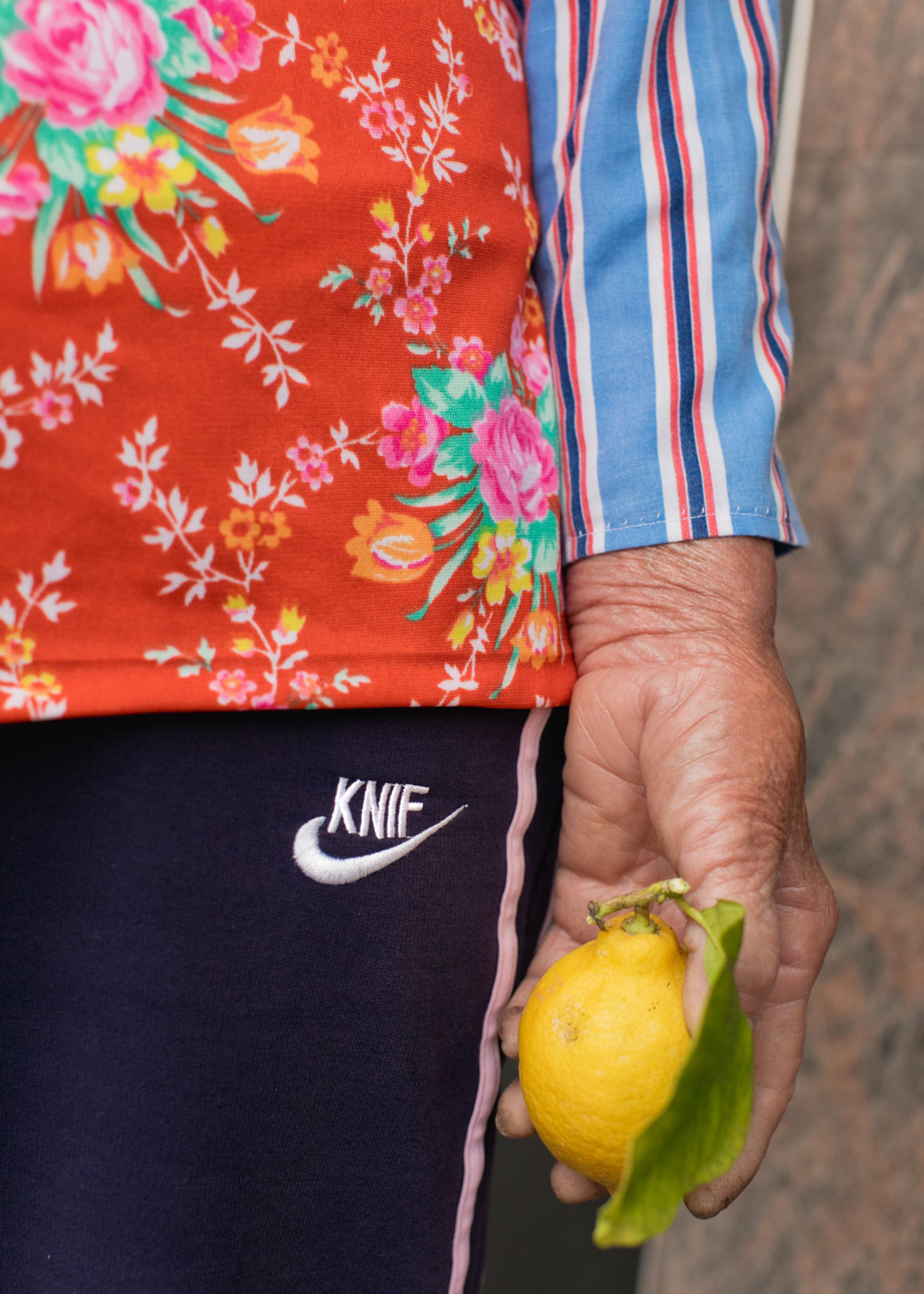A person wearing colorful floral and striped clothing holds a lemon with a green leaf in their hand, standing against a dark background.
