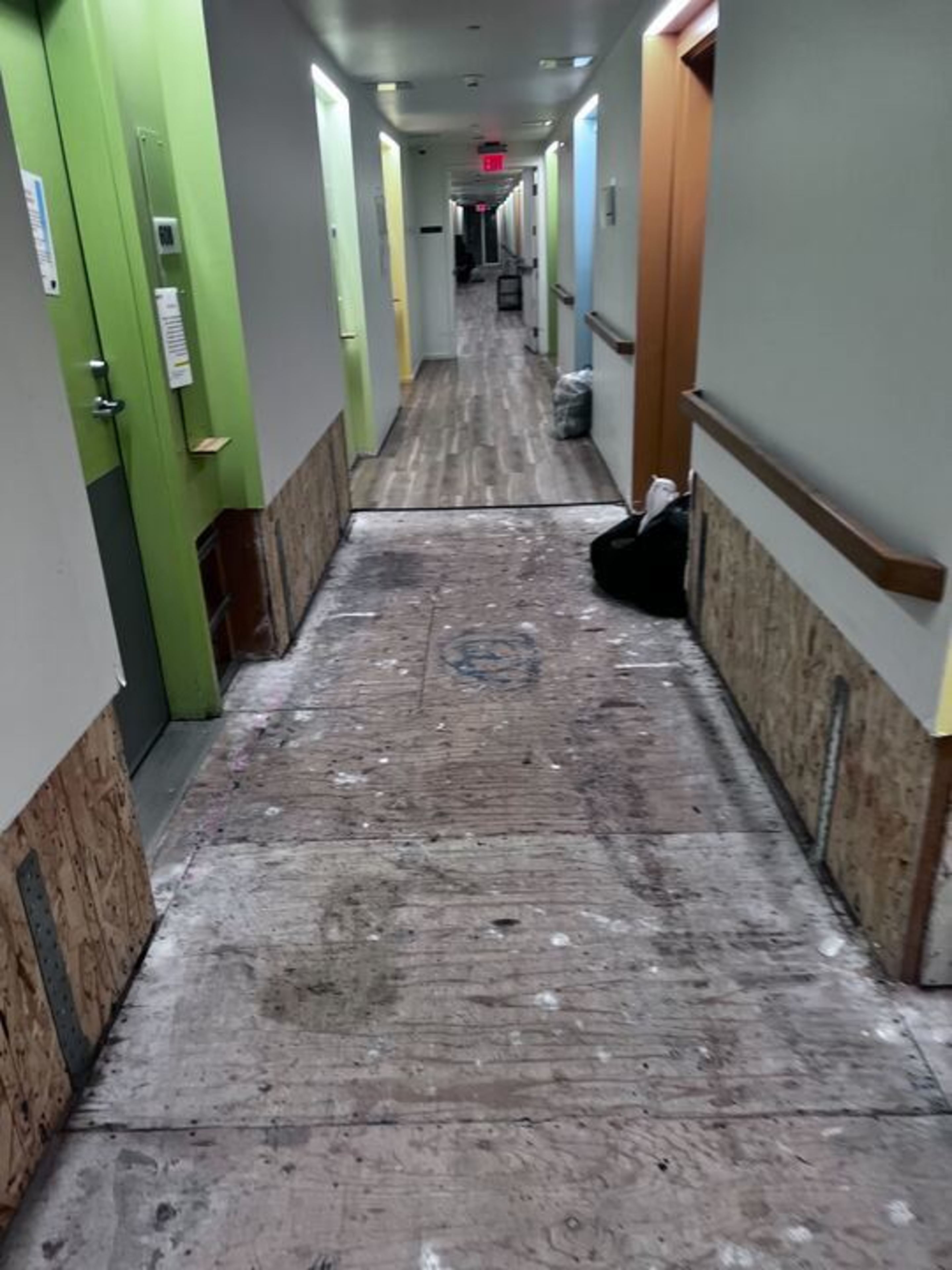 A long hallway with partially unfinished flooring, doors on both sides painted in green, yellow, blue, and orange, and walls lined with plywood.