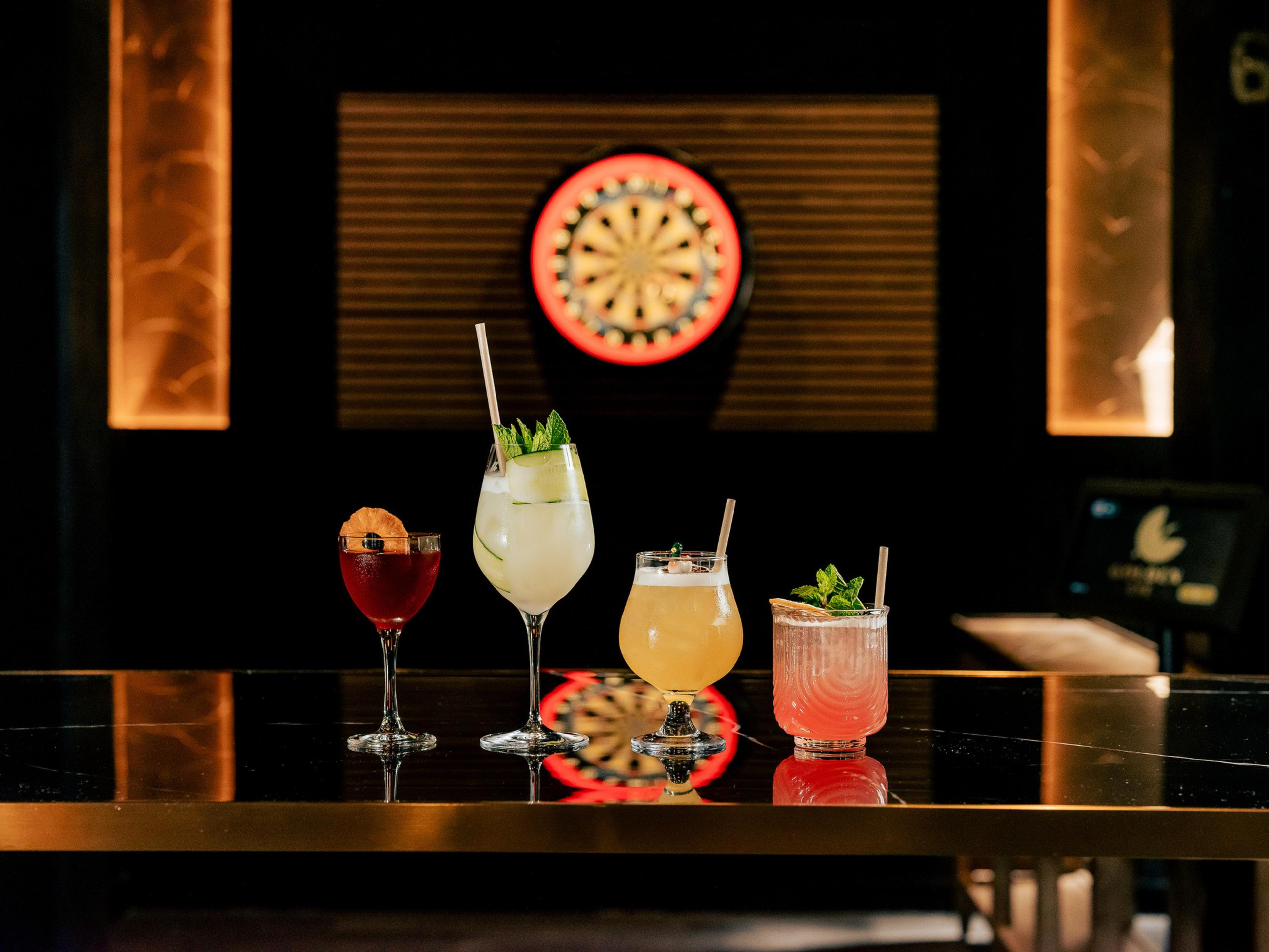 Four colorful cocktails with garnishes are lined up on a reflective table in a dimly lit room; a dartboard is visible in the background.