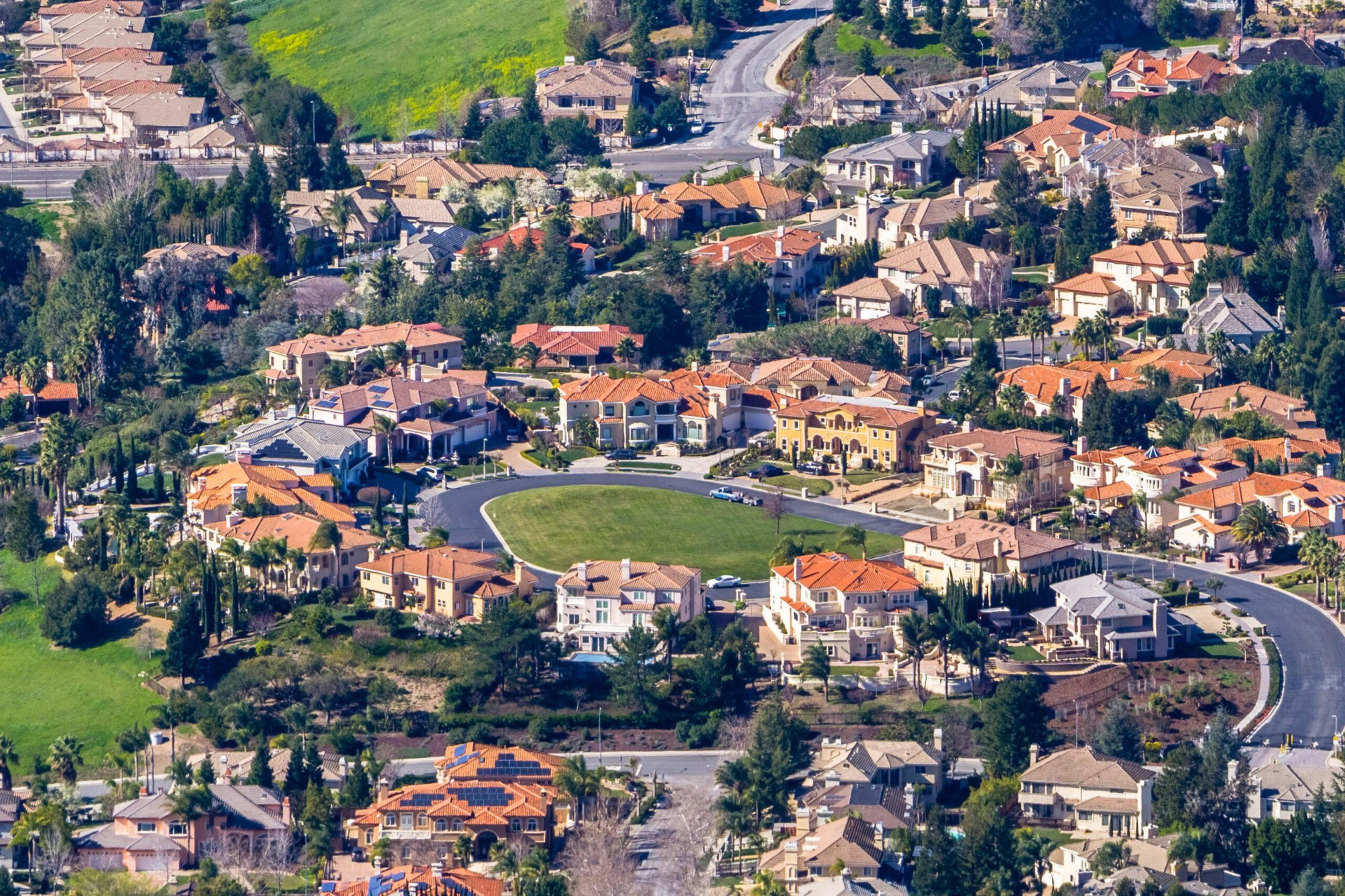 A spacious suburban neighborhood with large homes, red-tile roofs, green lawns, winding roads, and a central grassy oval area surrounded by houses.