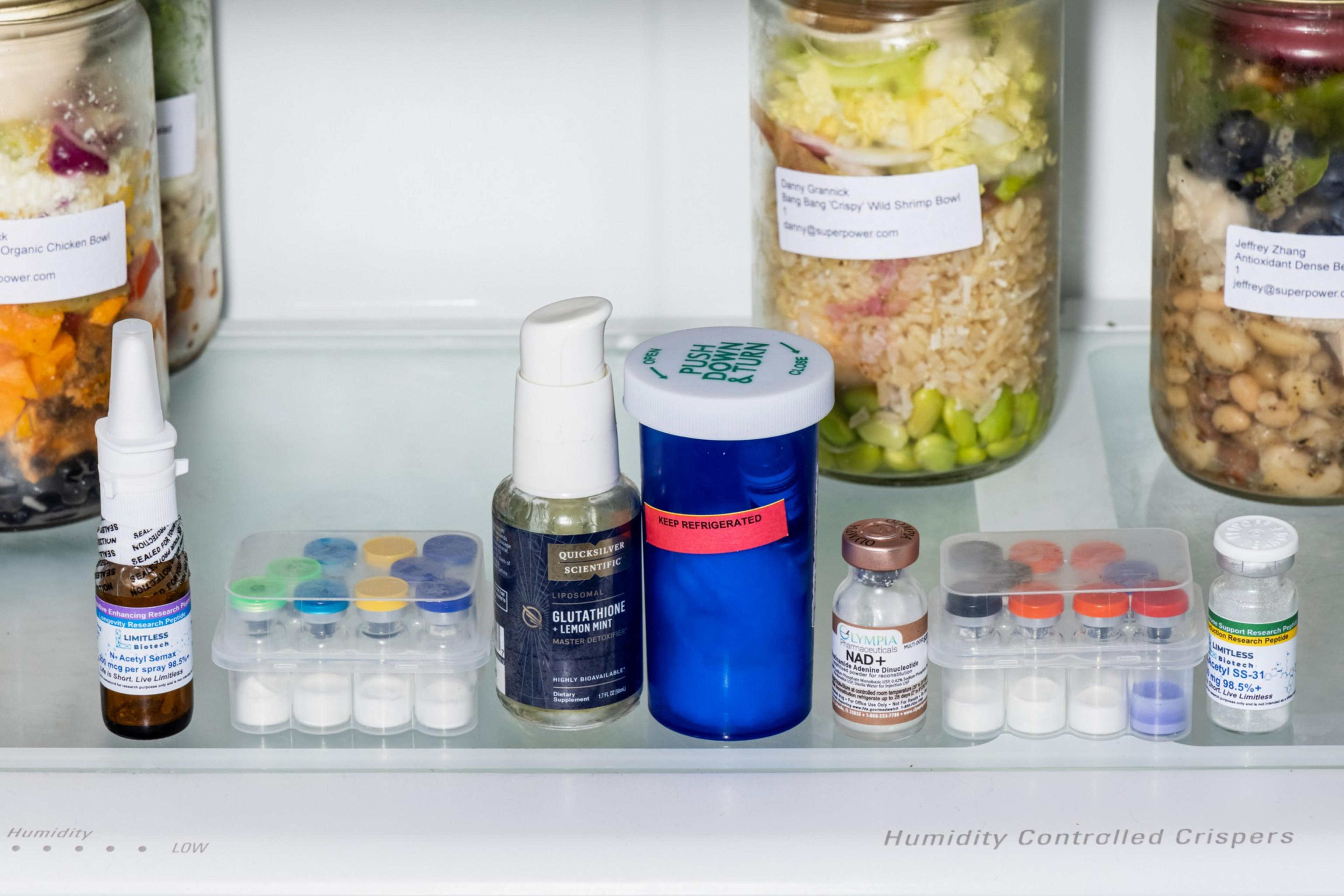 Several small medication bottles and vials are neatly arranged on a refrigerator shelf, with layered jars of colorful food in the background.