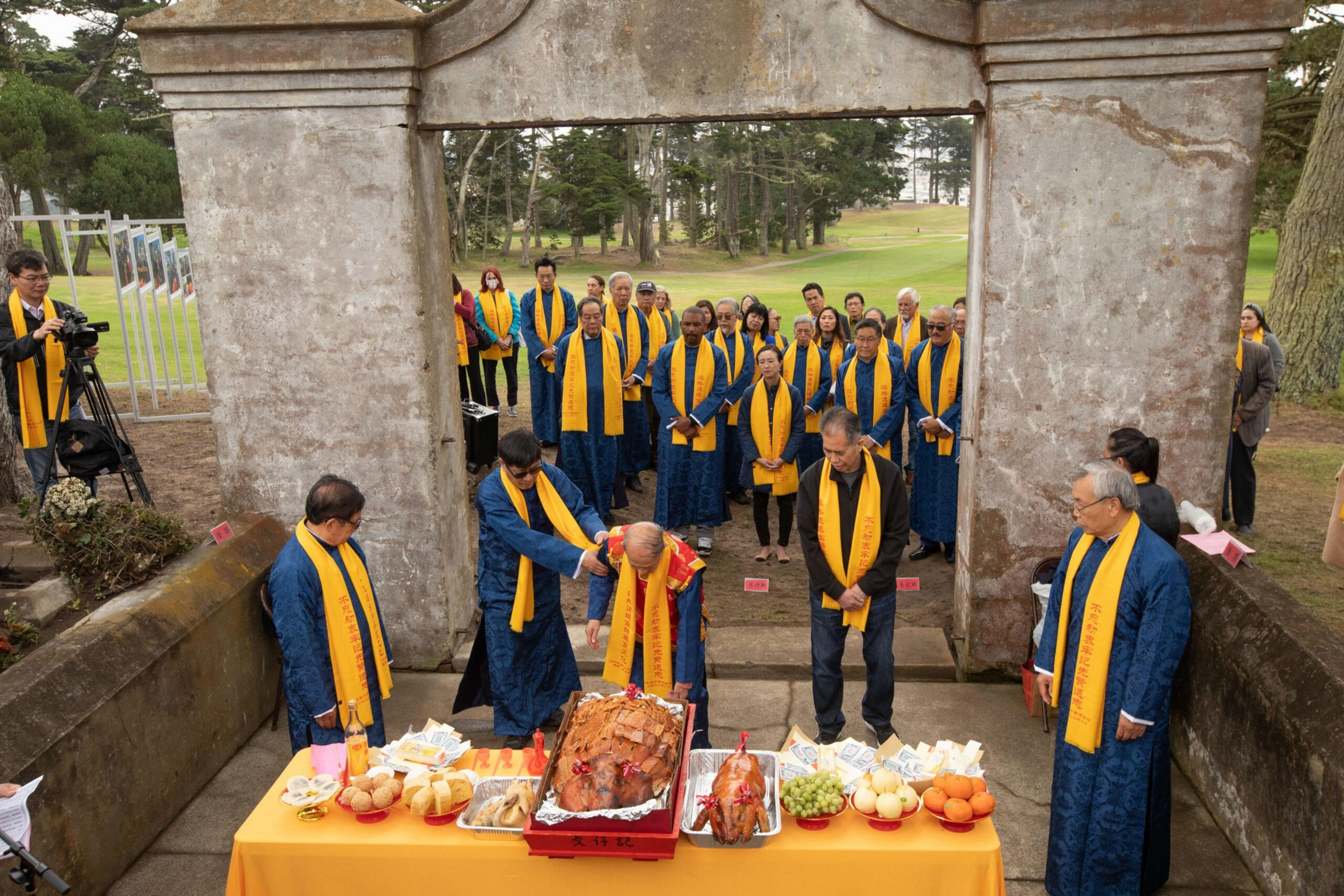 A group of people wearing blue robes and yellow scarves stand solemnly around a table with roasted pigs and offerings under a stone archway outdoors.