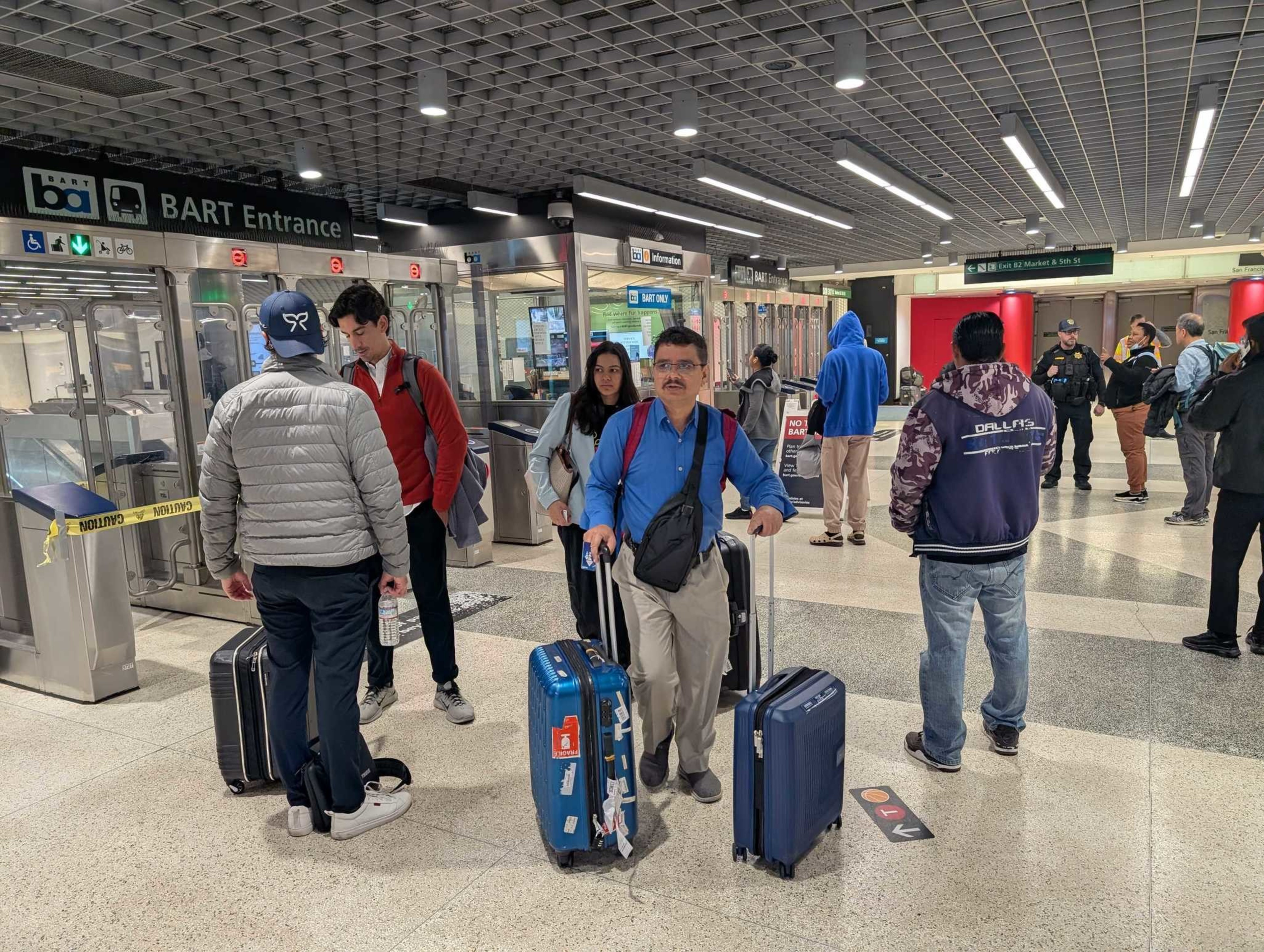 The image shows people with luggage inside a BART station entrance, surrounded by closed gates and caution tape. Some individuals are talking, and others are walking.