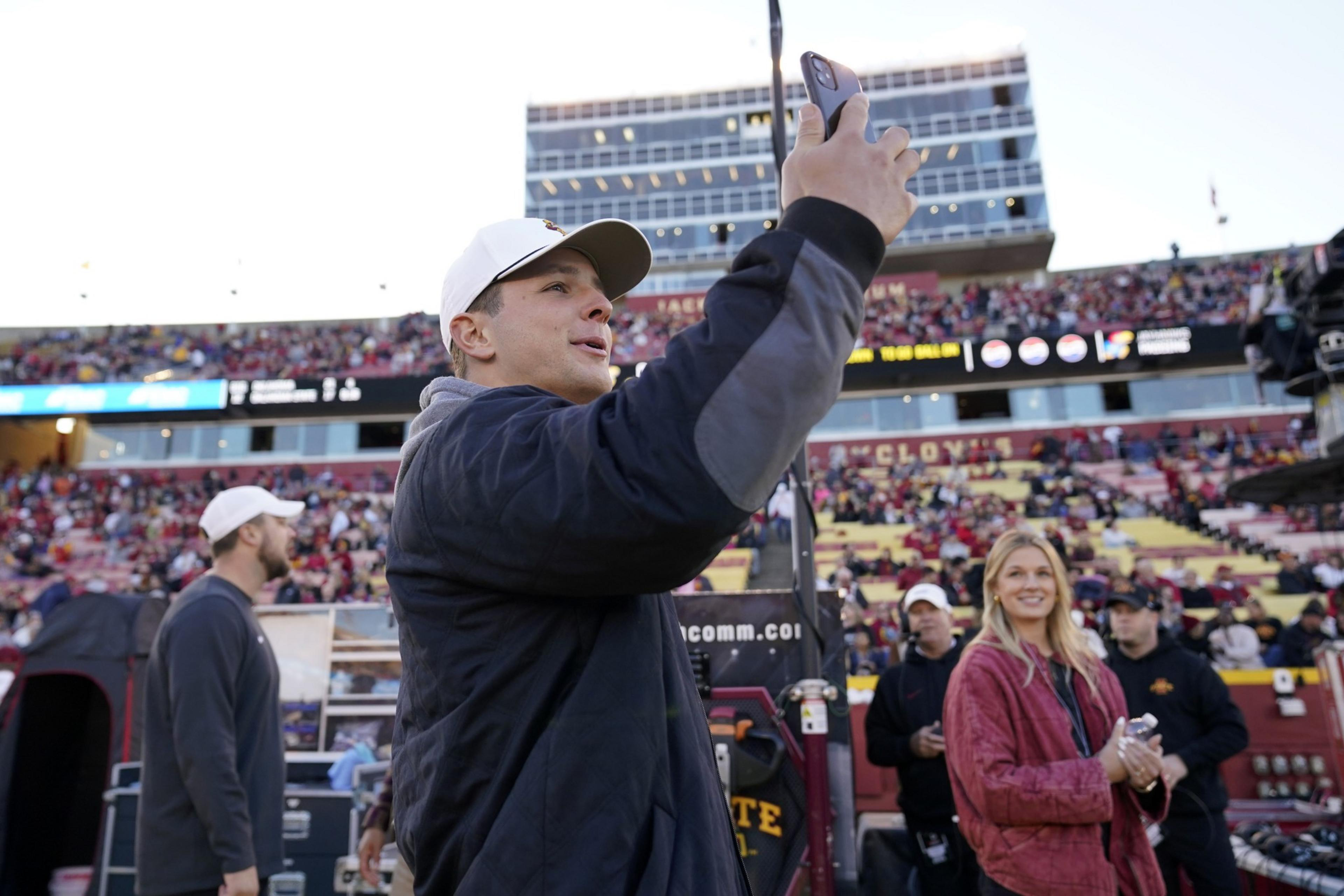 A man in a white cap takes a selfie on the sidelines of a sports stadium. A woman in a red jacket stands nearby, with a packed crowd in the background.