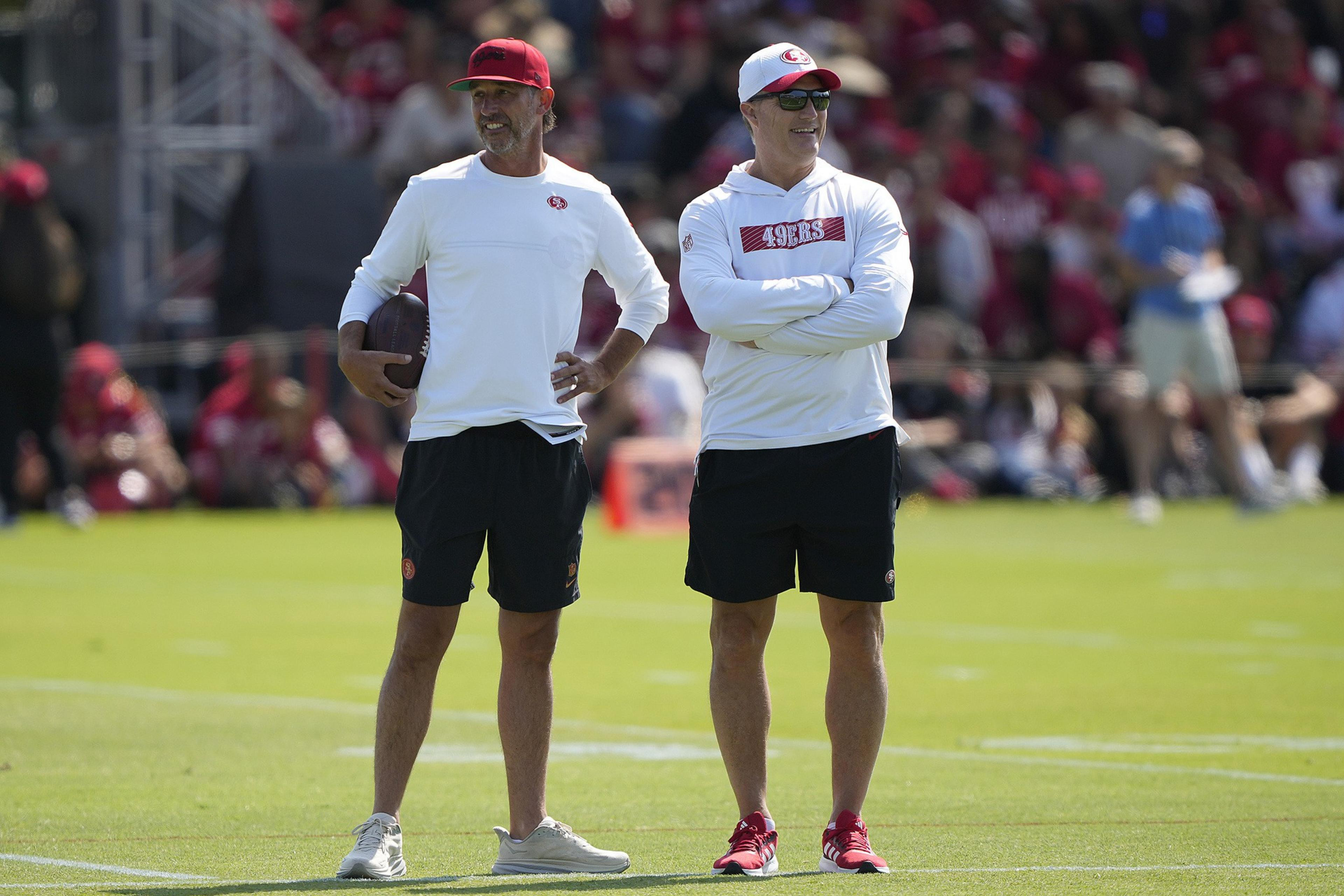 Two men in athletic attire stand on a field. One has a red cap and holds a football, while the other wears sunglasses and a white hat. A crowd is in the background.