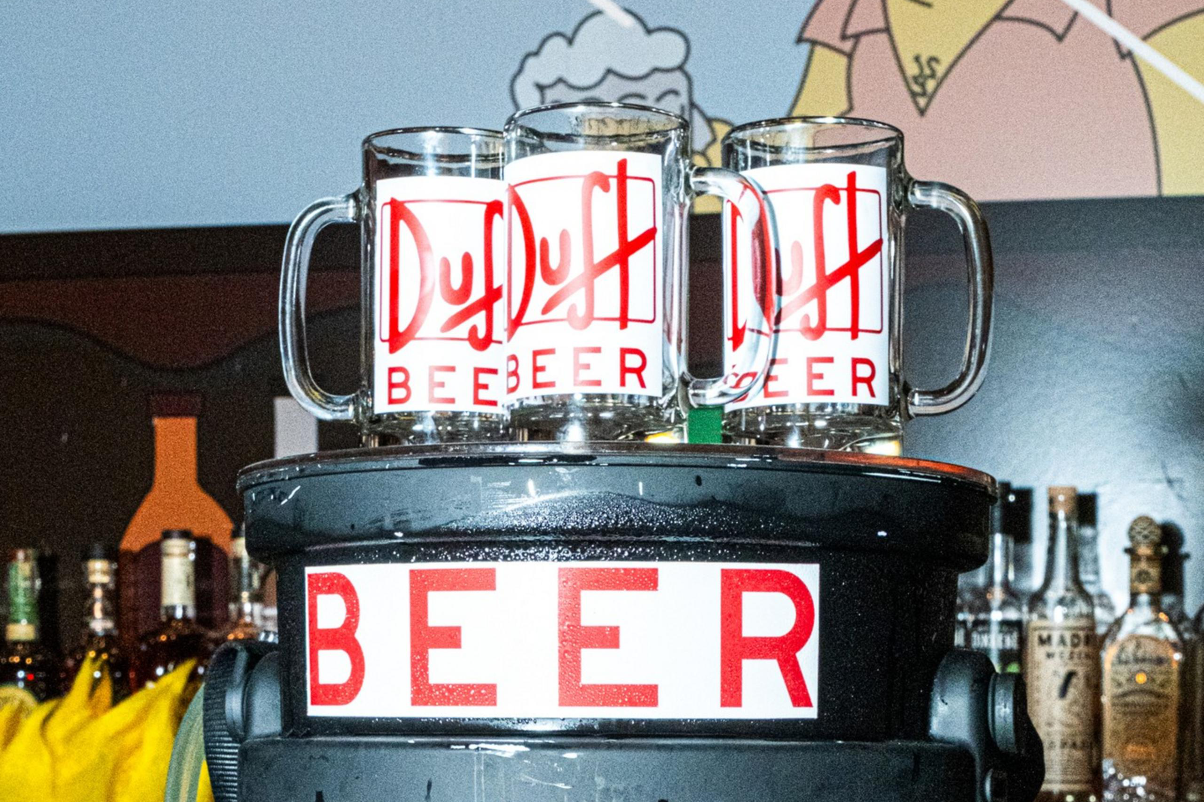 The image shows a bar setup with a large container labeled "Duff Beer" topped with two beer mugs. A cartoon character is on the wall behind.