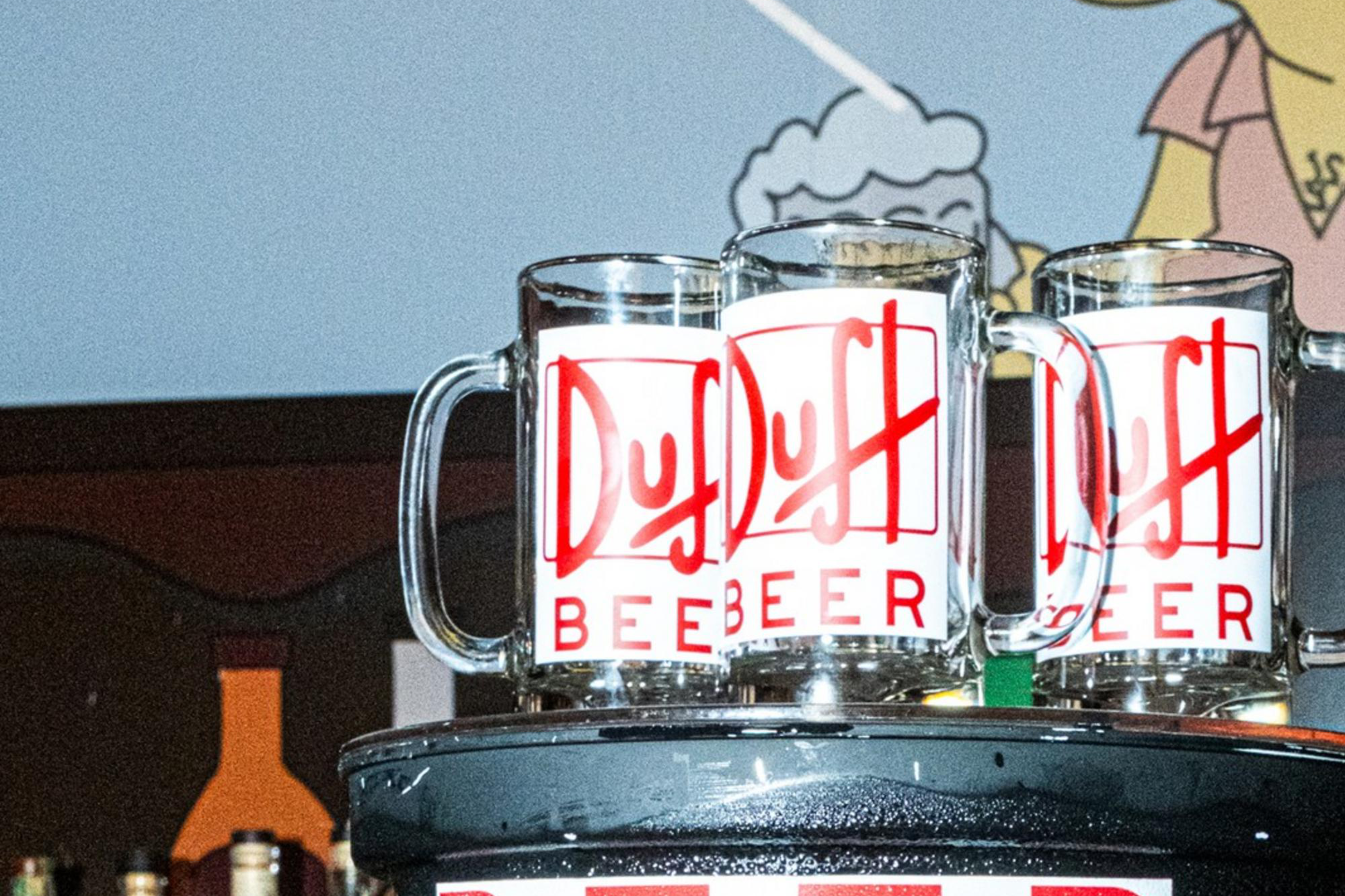 The image shows a bar setup with a large container labeled "Duff Beer" topped with two beer mugs. A cartoon character is on the wall behind.