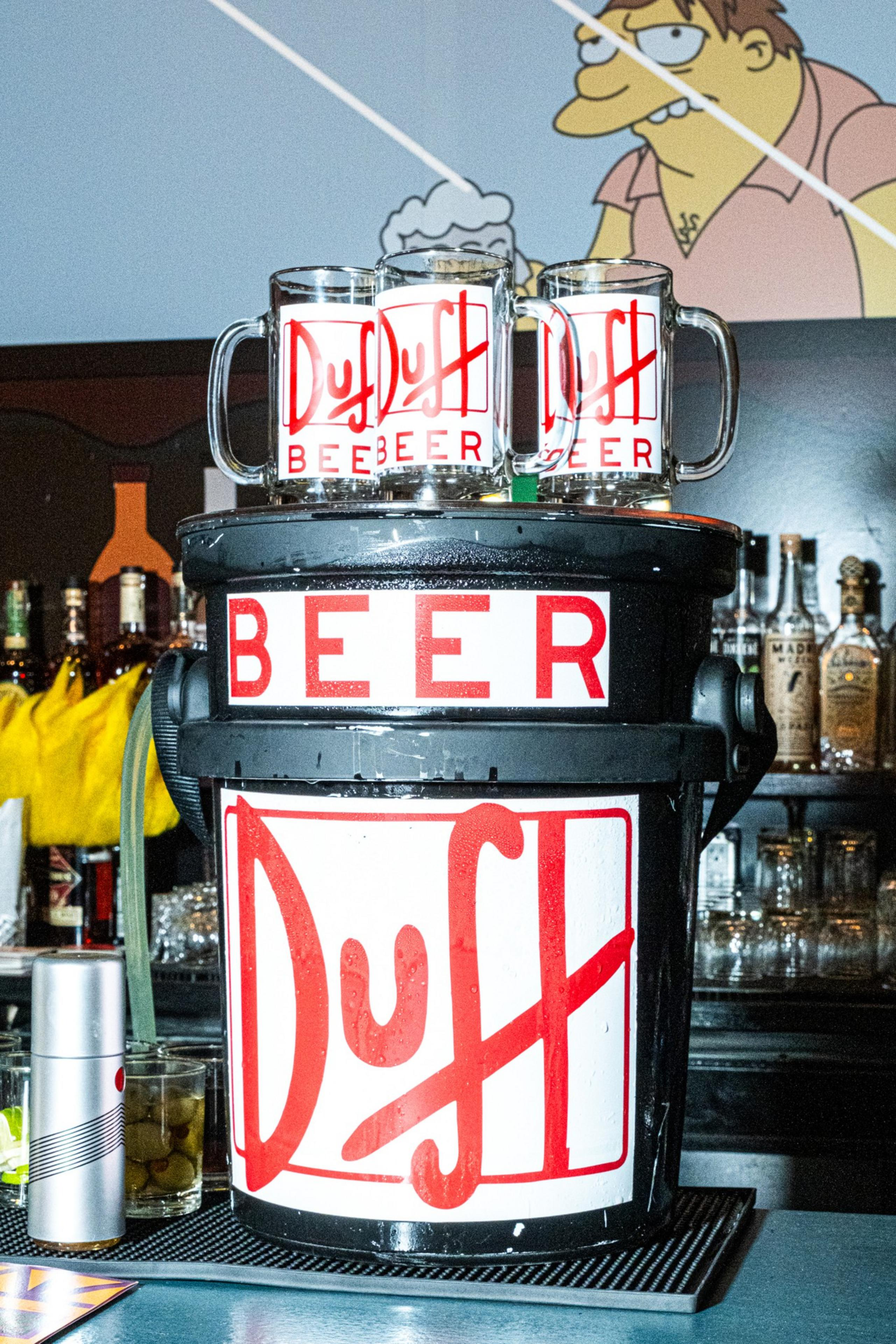 The image shows a bar setup with a large container labeled &quot;Duff Beer&quot; topped with two beer mugs. A cartoon character is on the wall behind.