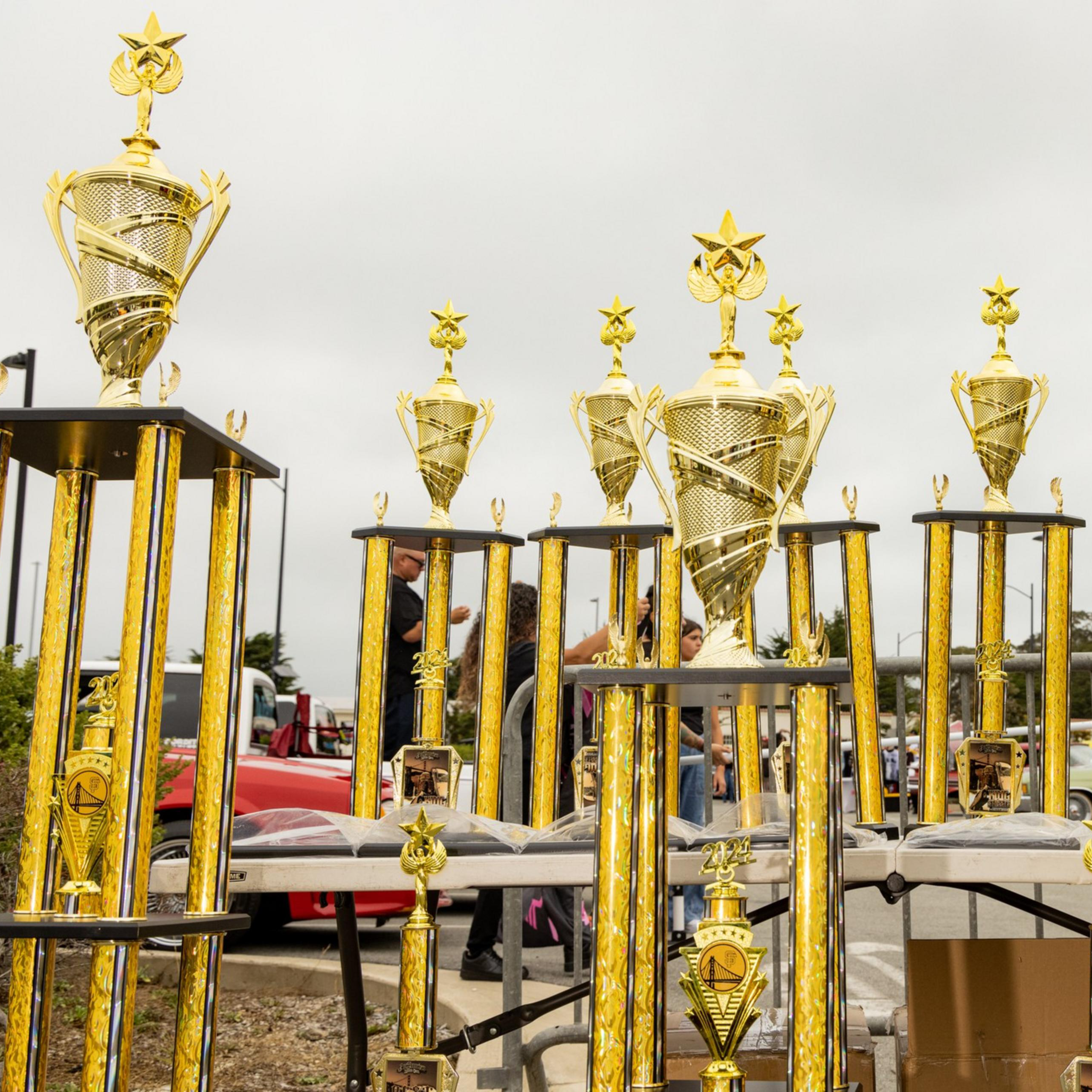 The image shows a variety of tall, golden trophies with intricate designs and star tops, displayed on tables outdoors with a cloudy sky in the background.