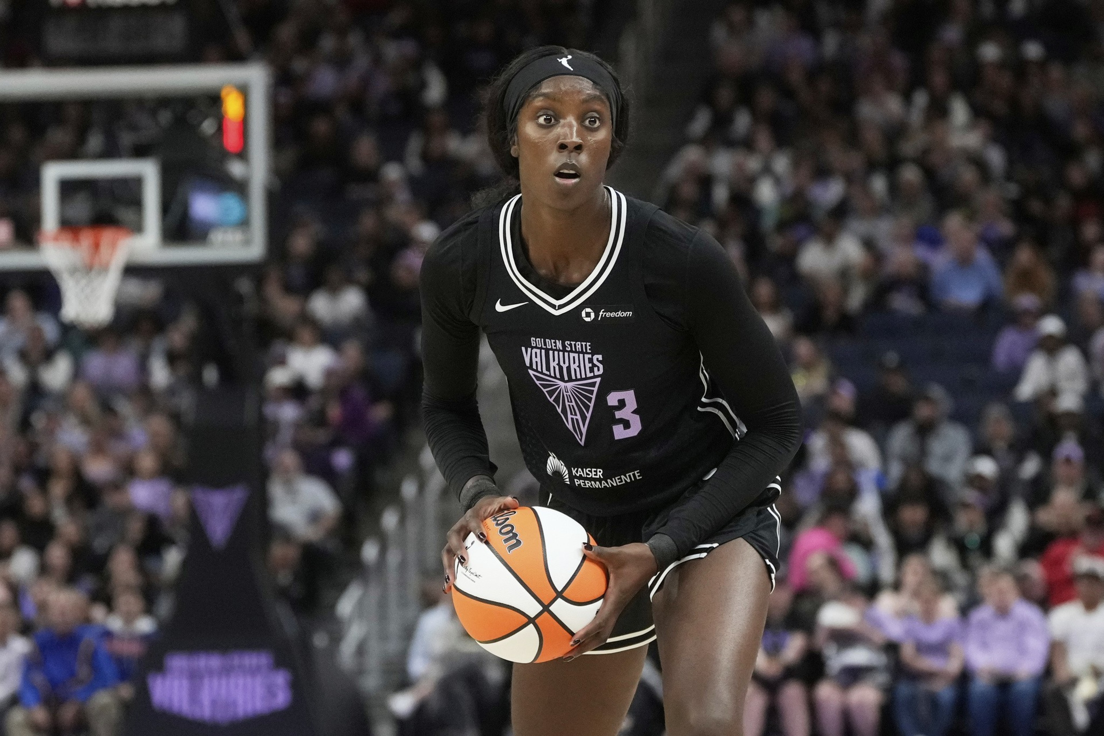 A basketball player in a black jersey is holding a basketball in a court. The crowd is blurred in the background, and the jersey reads &quot;Golden State Valkyries.&quot;