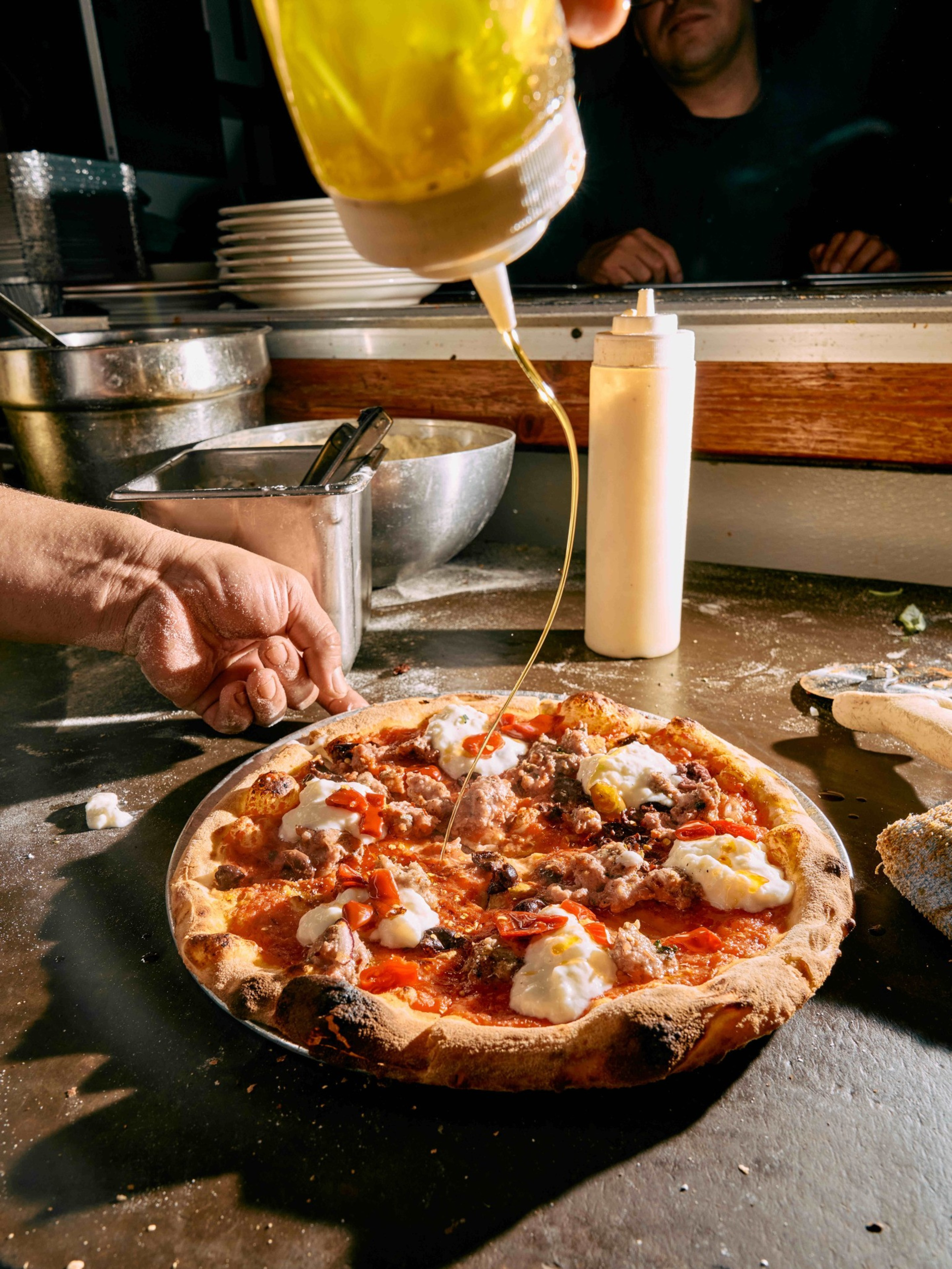 A hand pours olive oil from a squeeze bottle onto a freshly baked pizza topped with dollops of cheese, sausage, and roasted red peppers on a metal table.