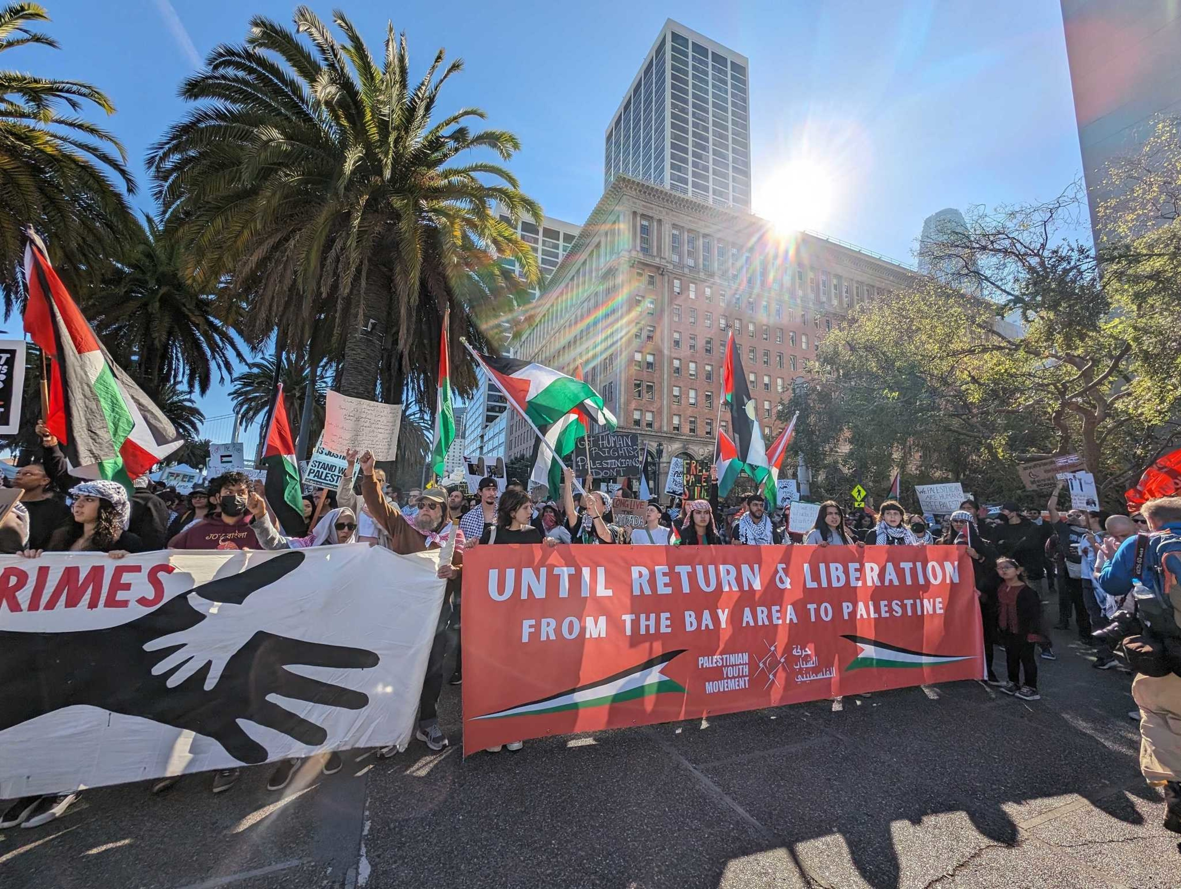 Dozens of people gather behind a red banner and wave black, green, red and white flags.