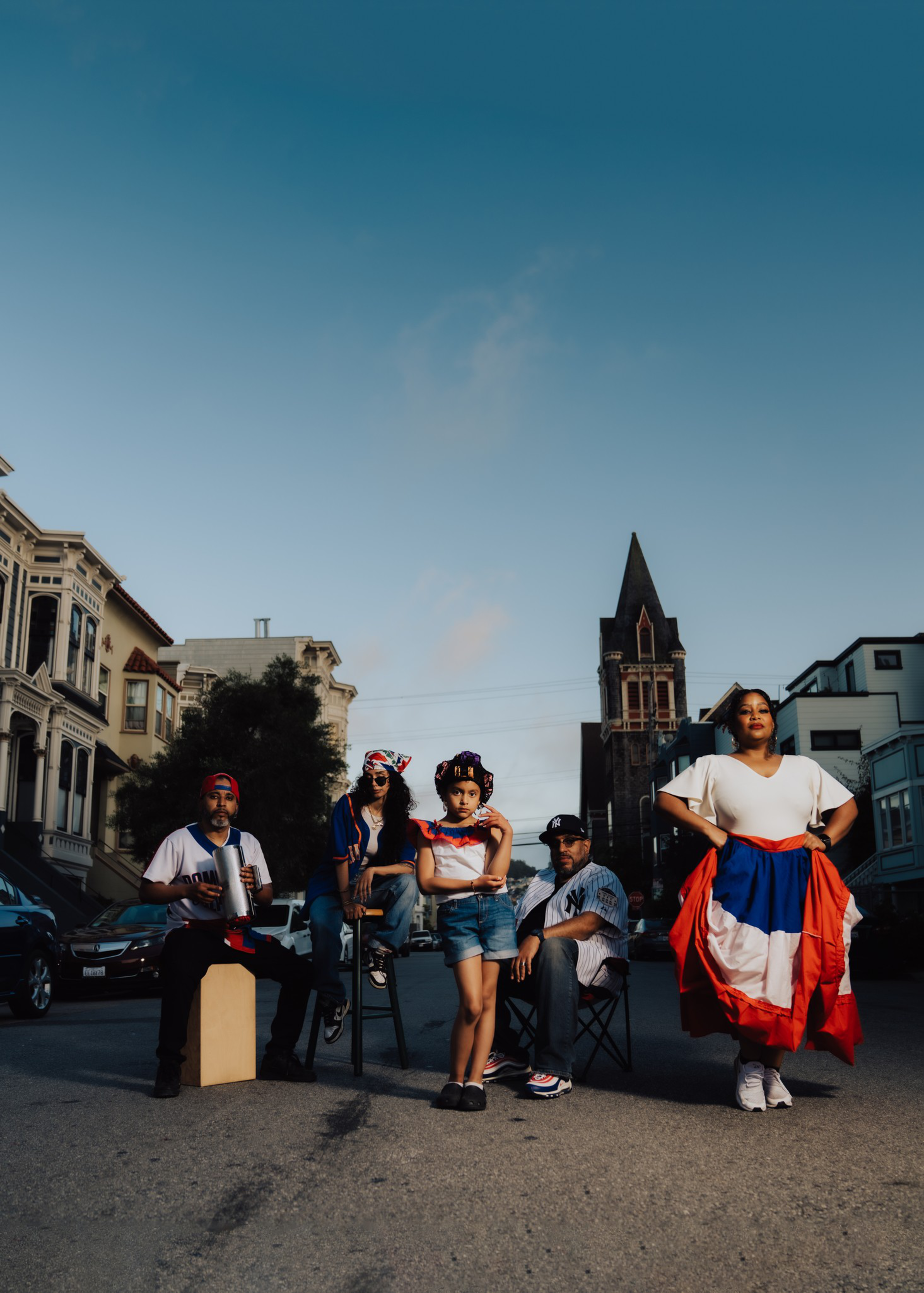 Five people of different ages stand and sit confidently in the middle of a street, some wearing clothing with red, white, and blue colors.