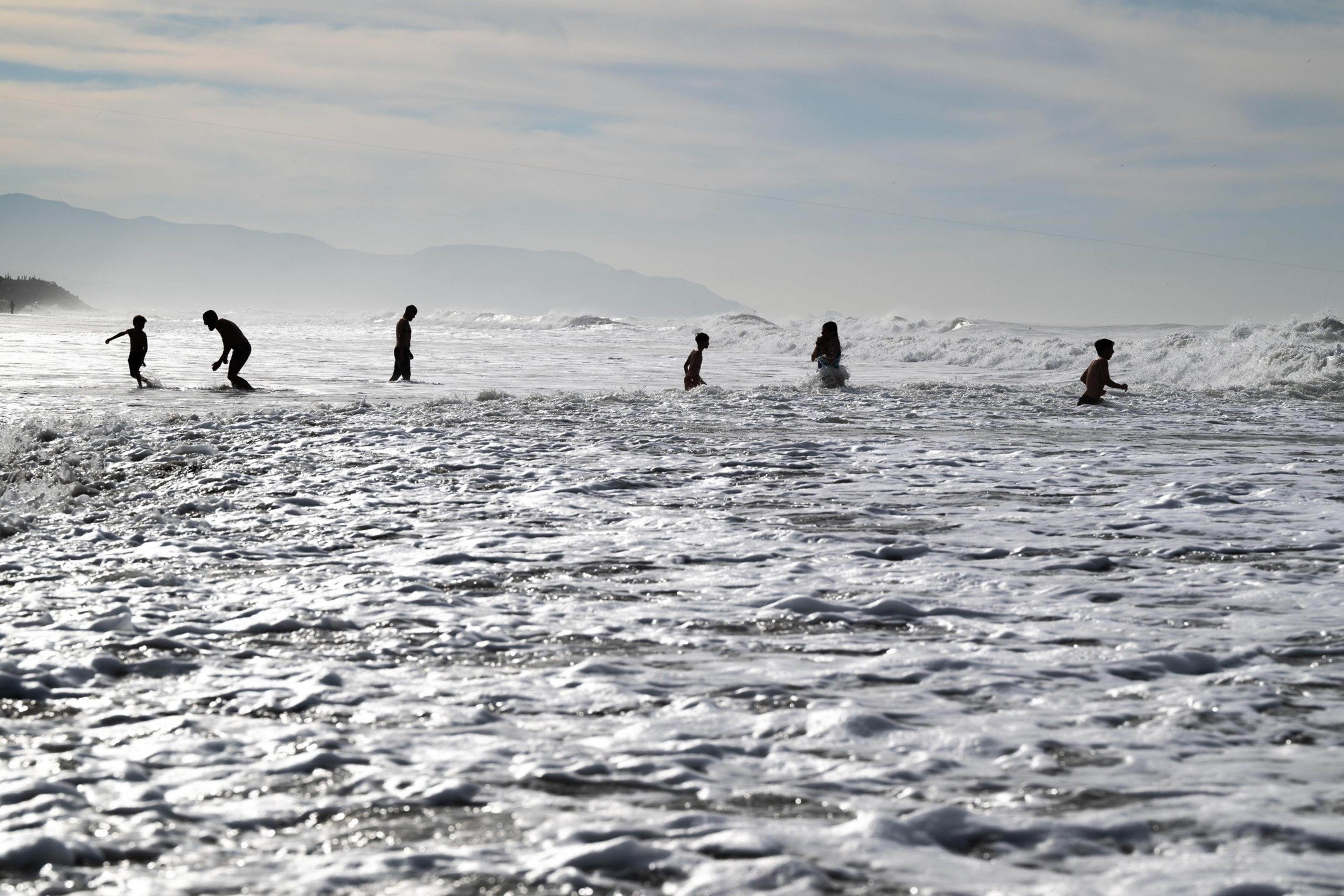 Several people are playing in the ocean's surf with gentle waves. The scene is set against a distant backdrop of hazy mountains under a cloudy sky.