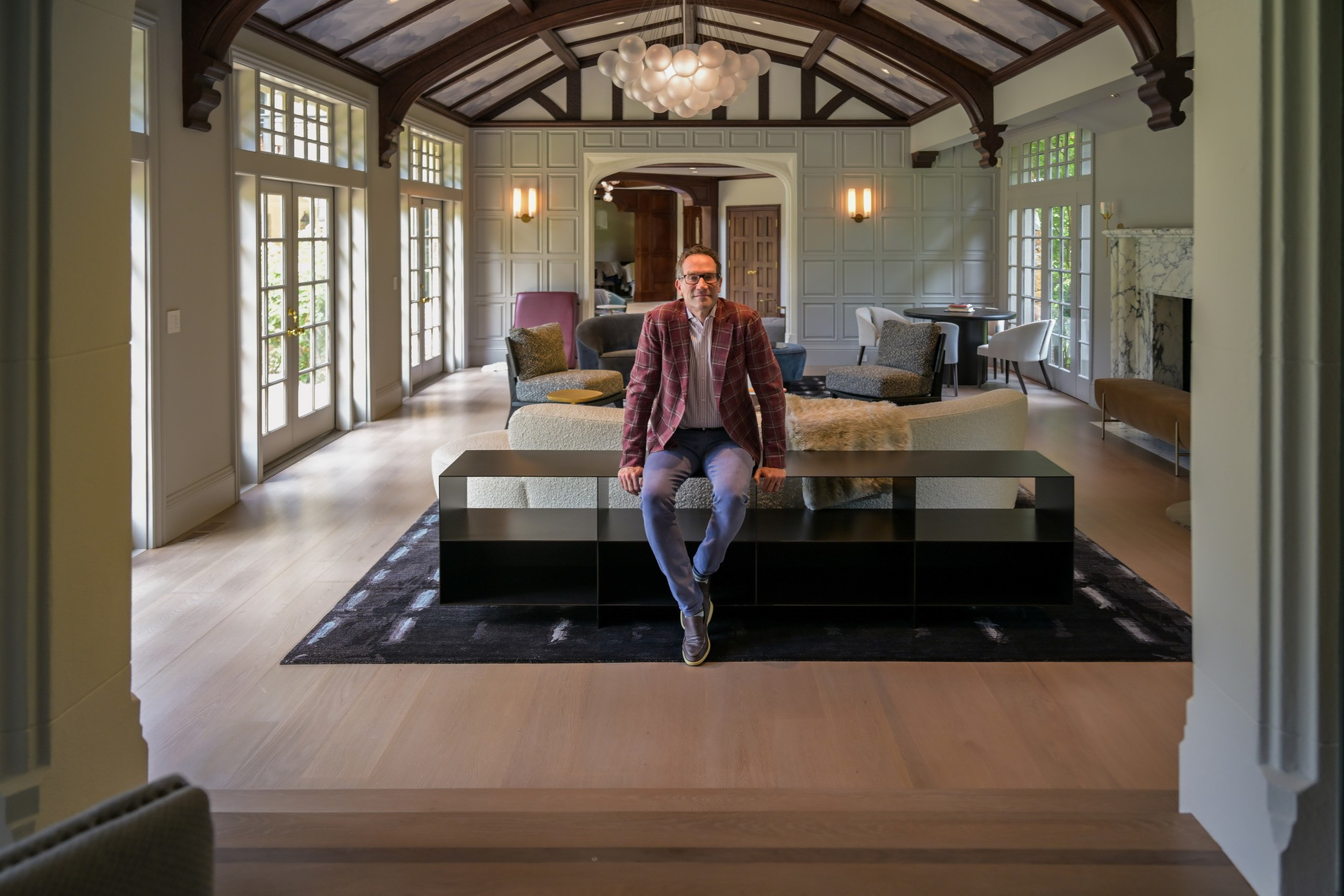 A man in a plaid jacket sits on a modern bench in a spacious, sunlit living room with high ceilings, large windows, and elegant furniture.